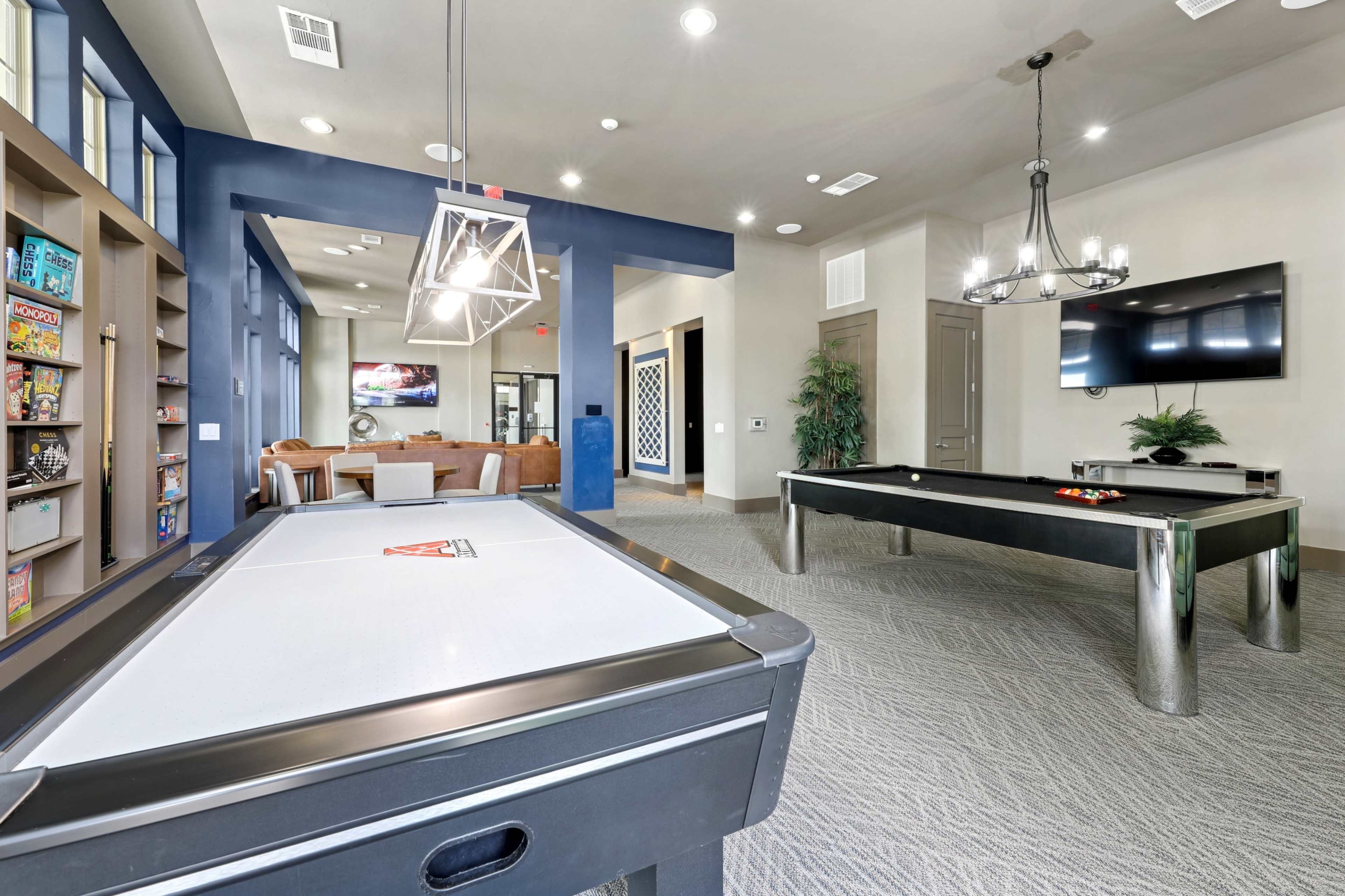 The image shows a recreational room featuring a pool table and a game table, with a seating area and shelving for board games in the background.