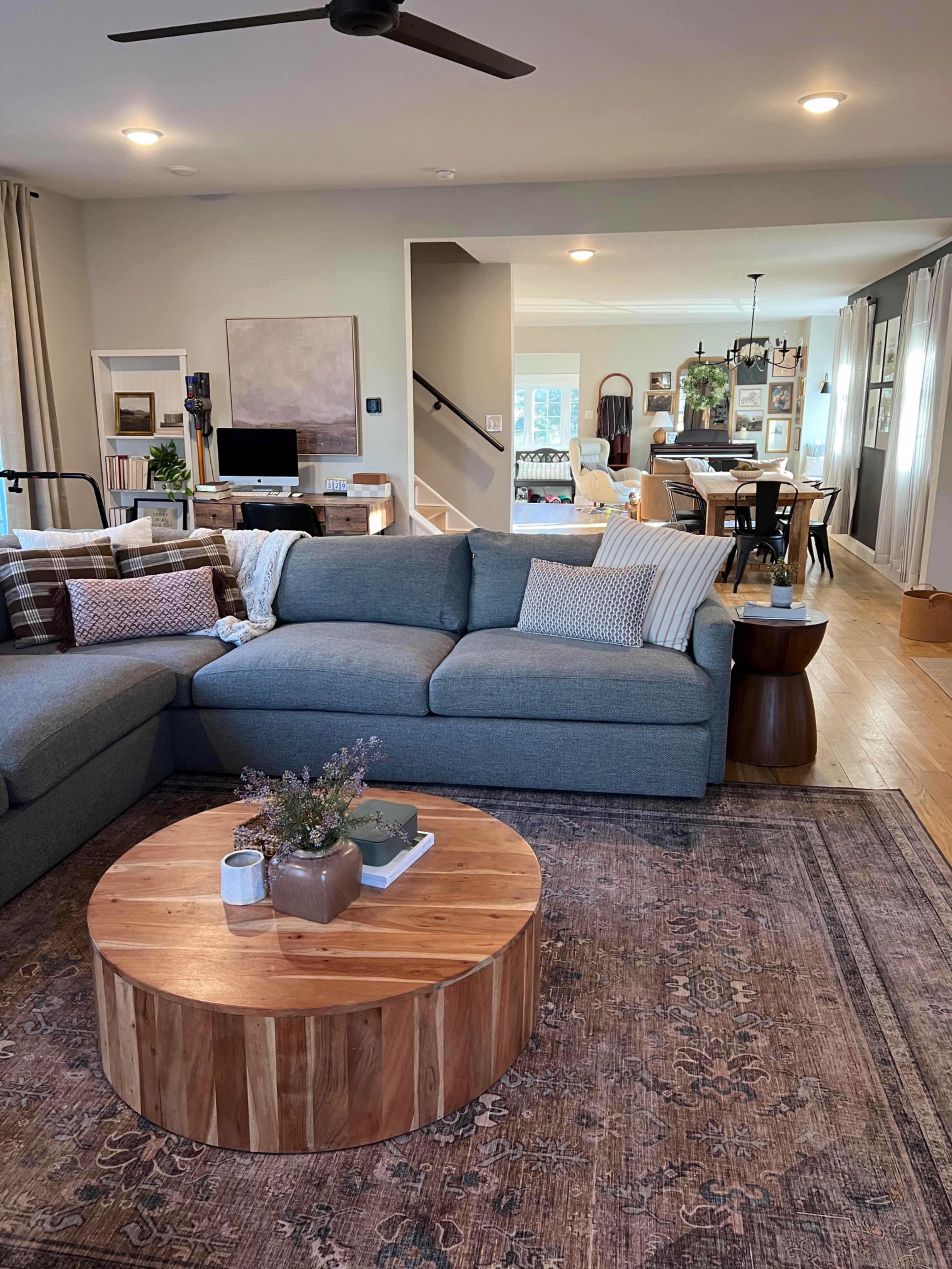 A spacious living room features a grey sectional sofa, a circular wooden coffee table, and an intricate area rug, leading into a dining area visible in the background.