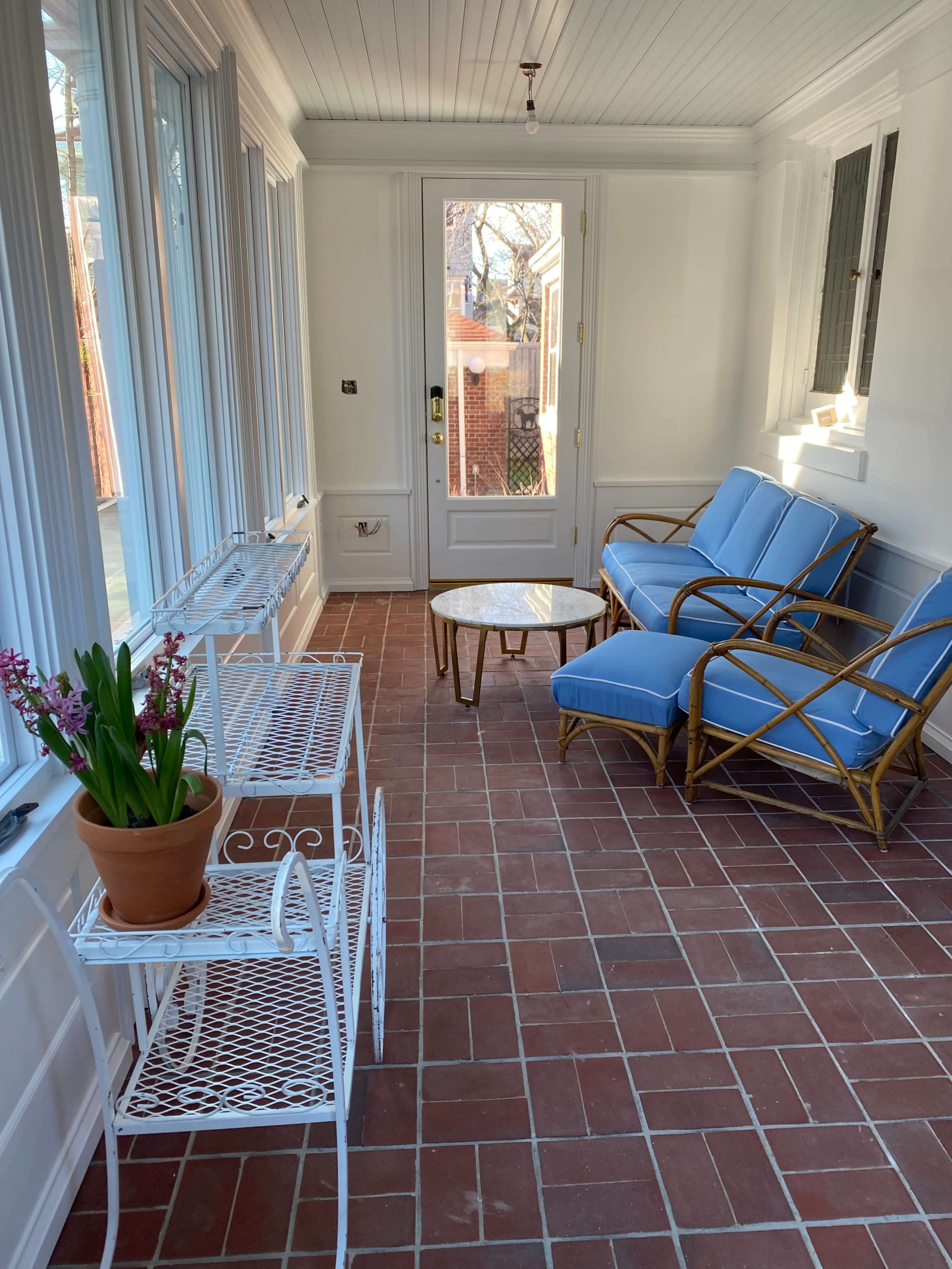 A bright sunroom features a small round table, a potted plant, and a blue seating arrangement against white walls and large windows.