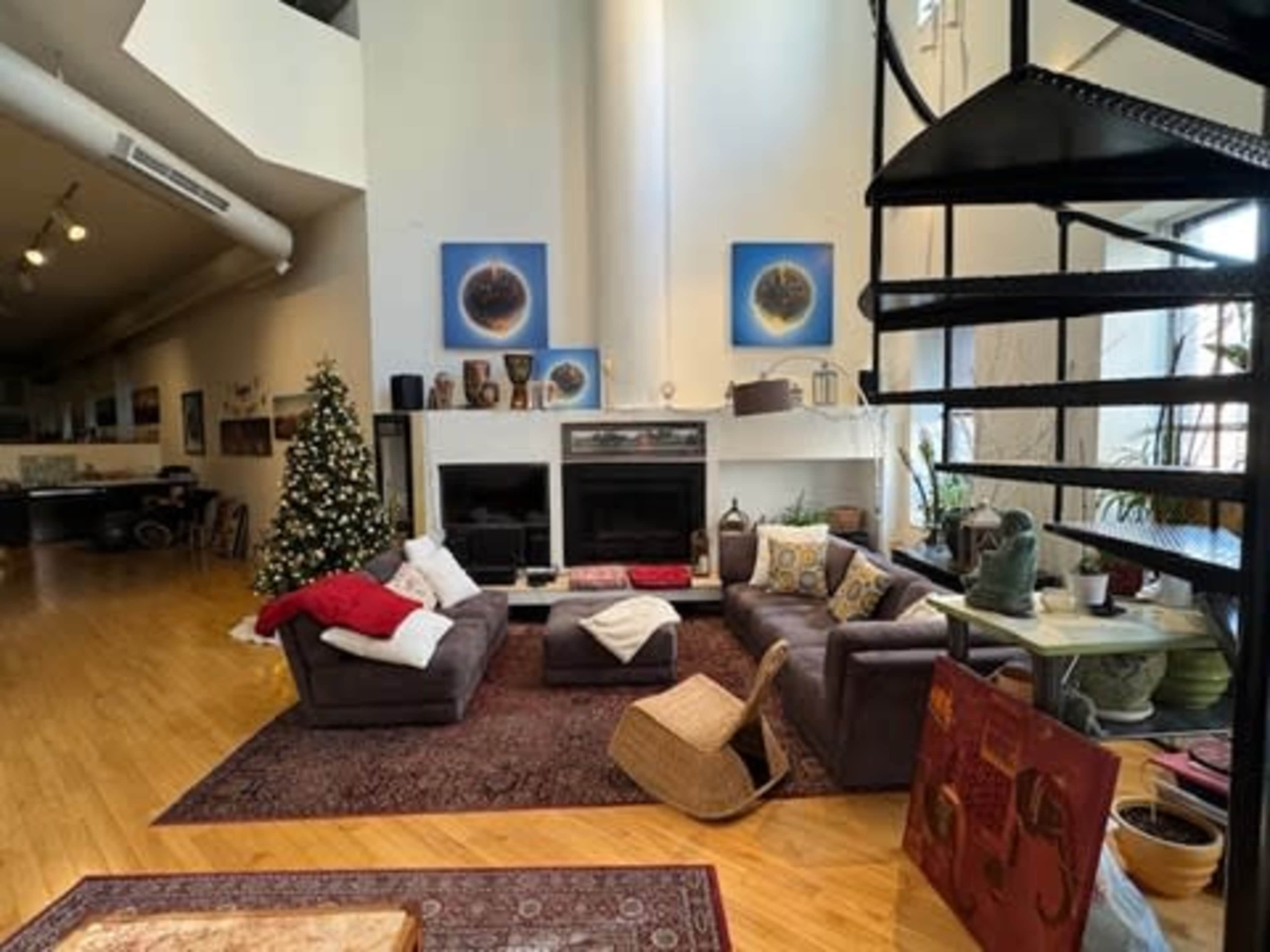 The image shows a spacious living room featuring a large sectional sofa, a coffee table, and a spiral staircase, with a Christmas tree in the corner and various artworks hanging on the walls.