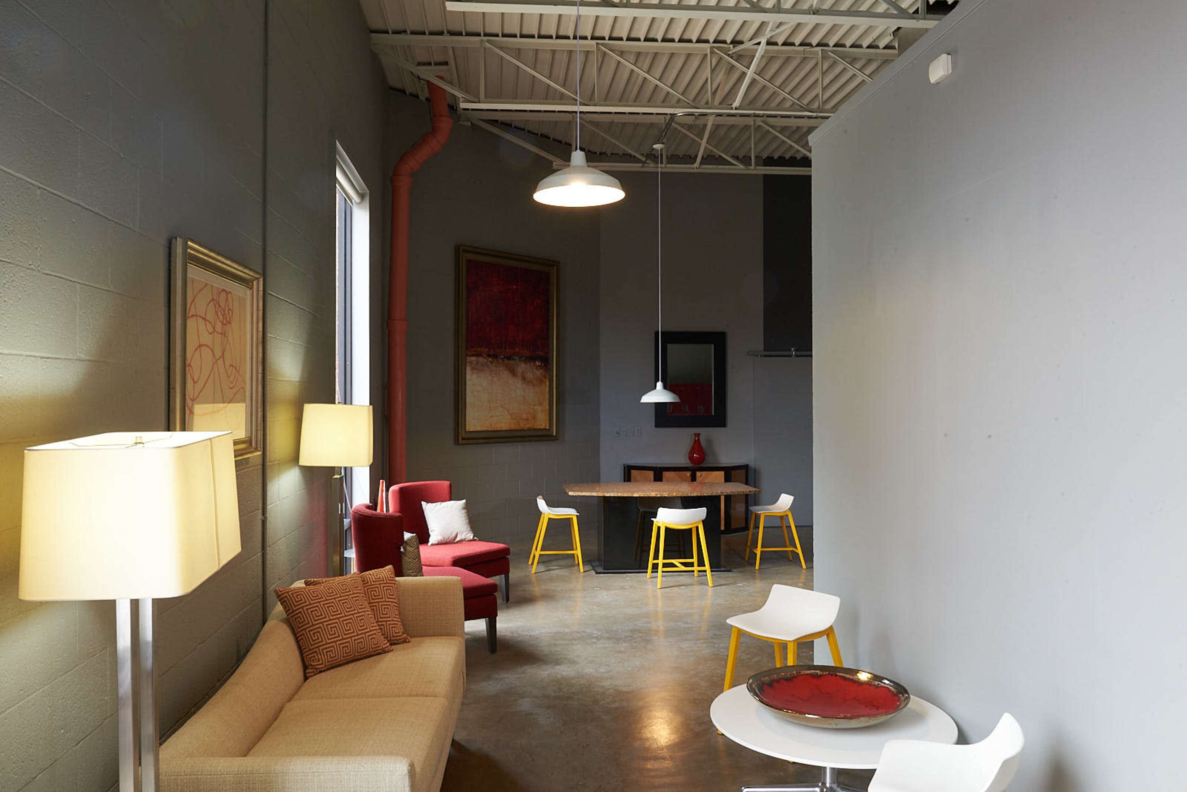A modern, open interior space featuring a beige sofa, a red armchair, a dining table with yellow chairs, and a lamp beside a wall with artwork.