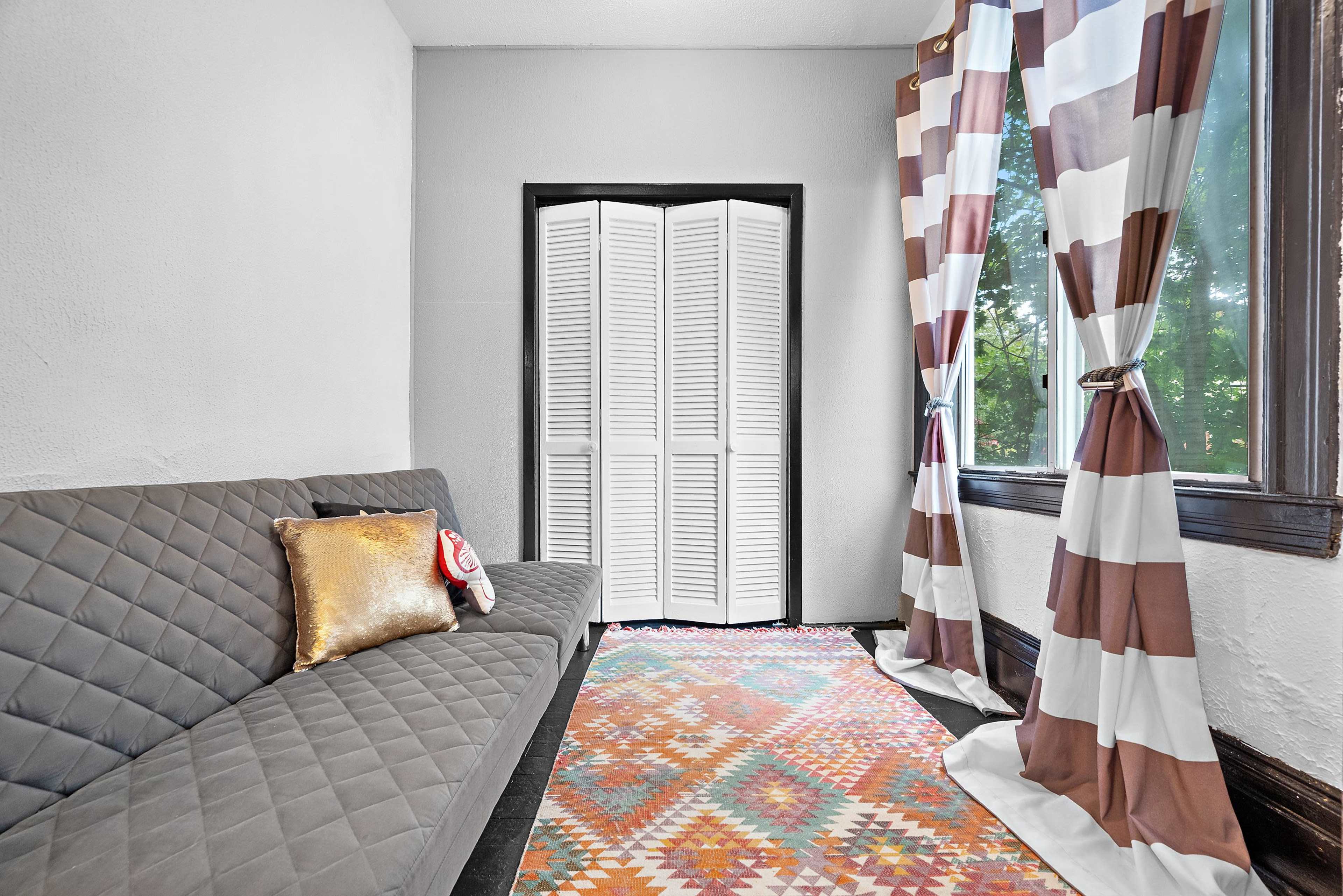 A small room features a quilted gray sofa, a colorful patterned rug, and striped curtains framing a window.