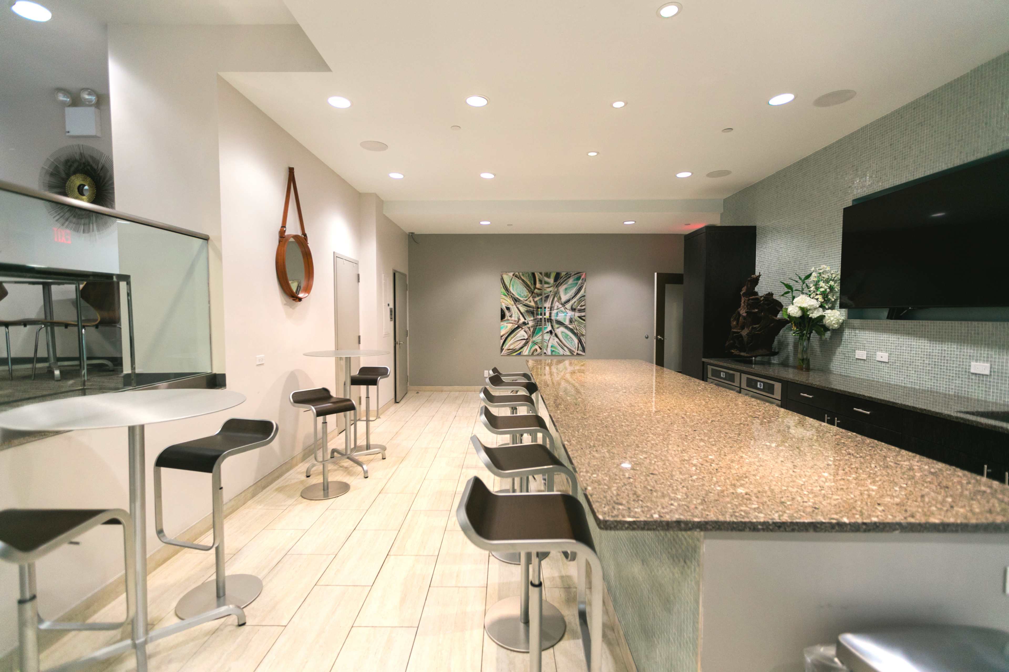 A modern bar area features a long granite countertop with high stools and minimalist decor, including an artwork and a television on the wall.