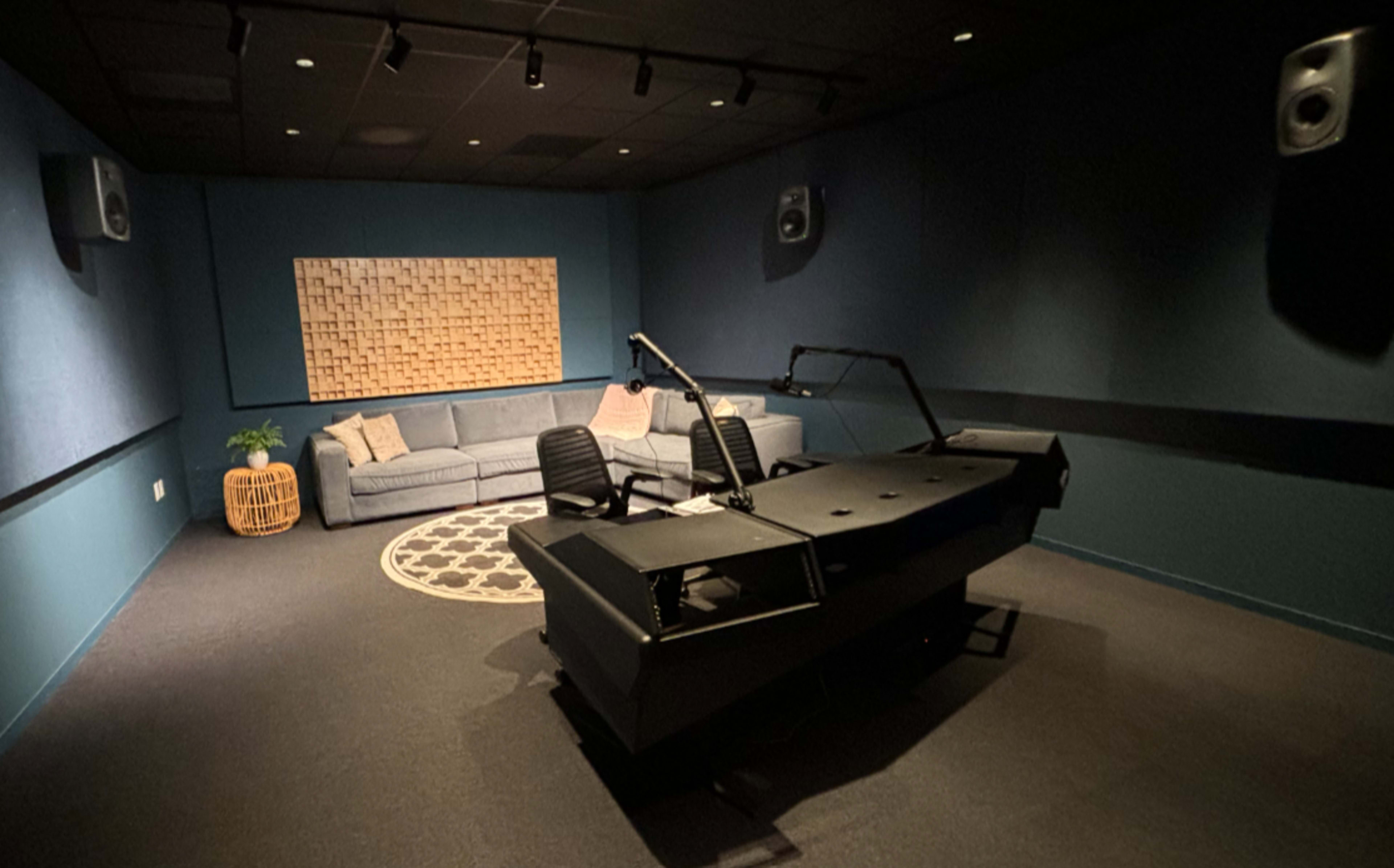 The image shows a modern recording studio with a console, two chairs at a desk, a sofa, and a wooden wall panel, surrounded by soundproofed walls.