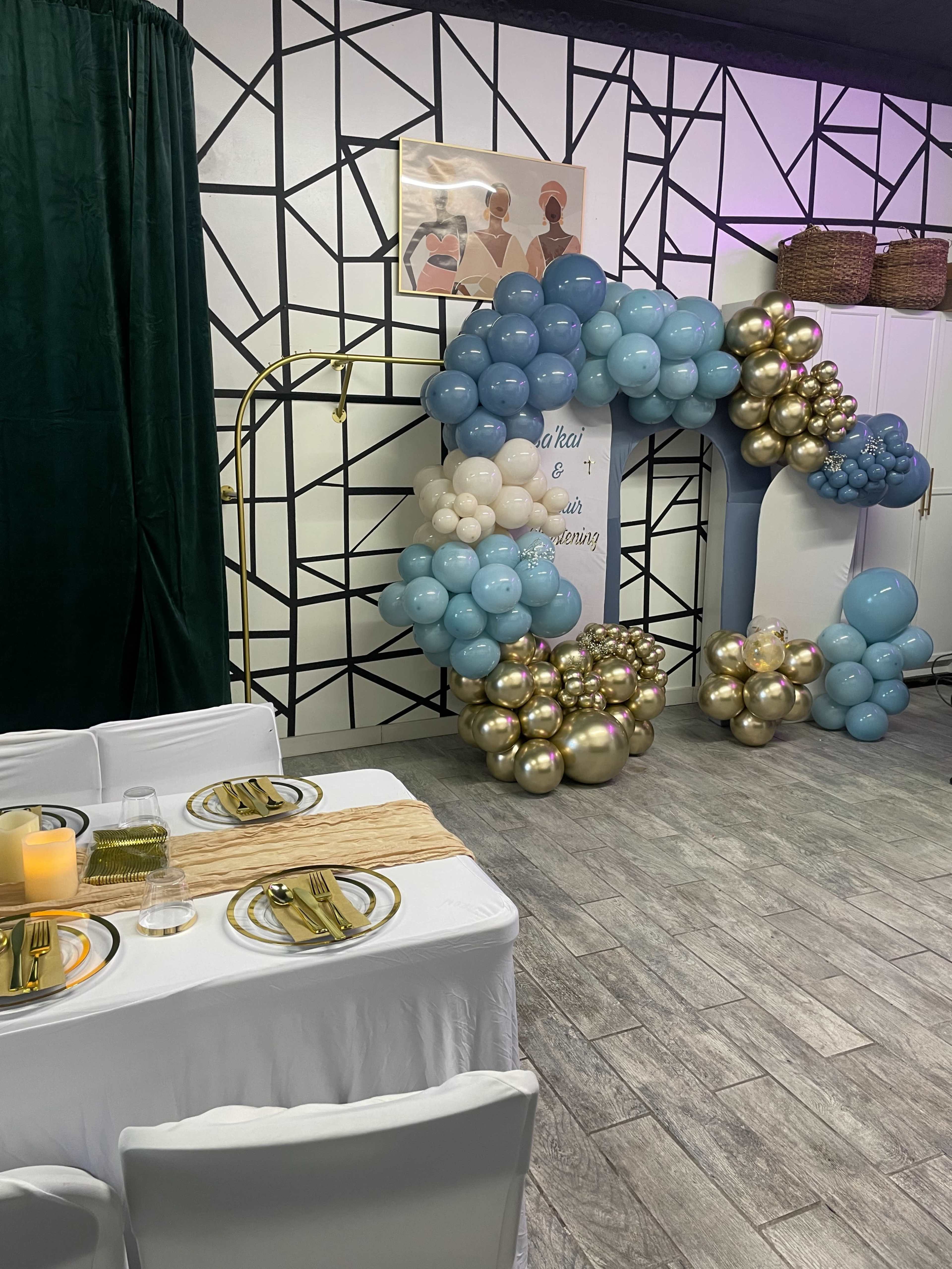 The image shows a decorated event space featuring a table set for dining, a balloon arch, and a wall with geometric designs.