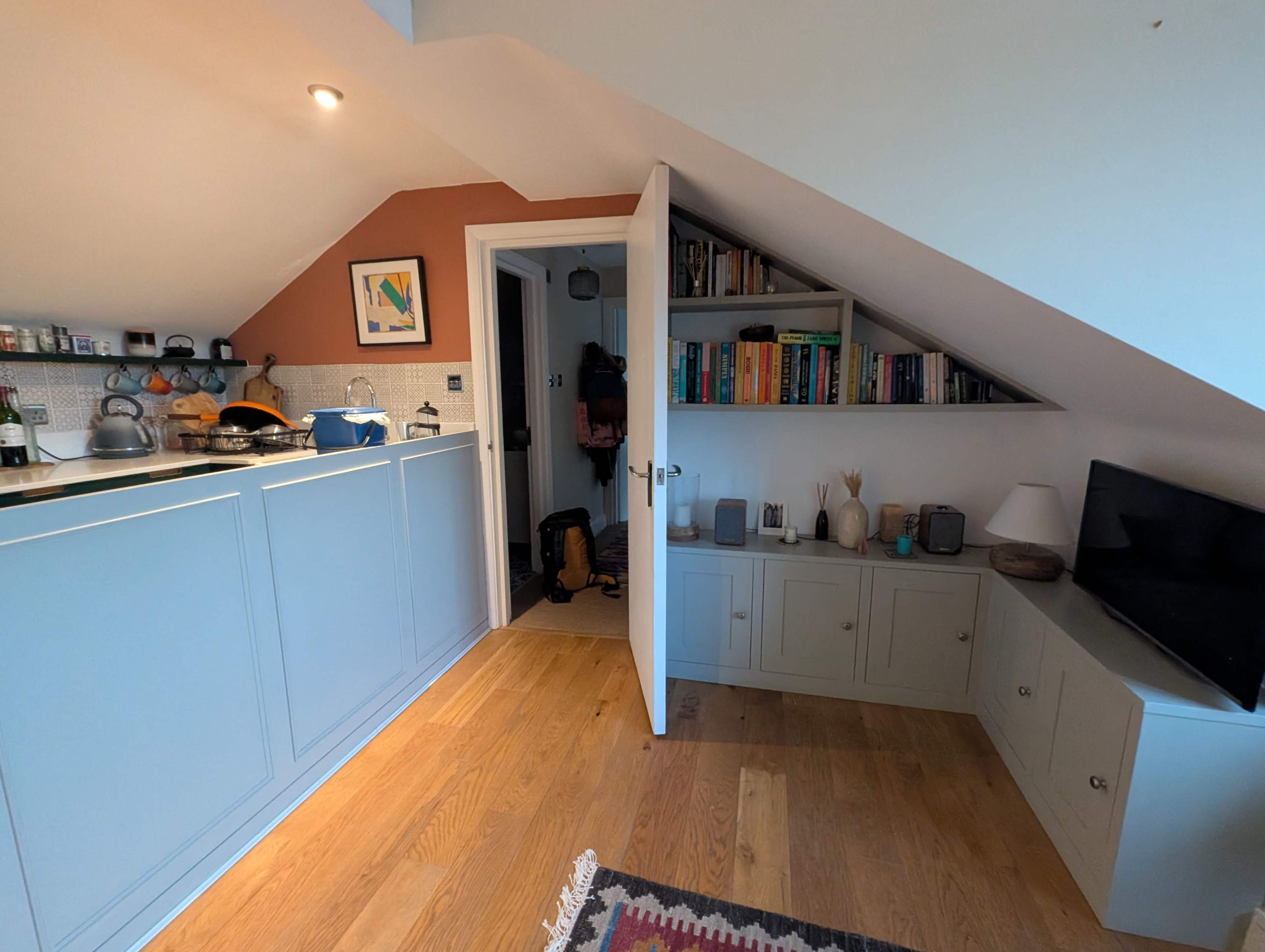 The image shows a small, angled room with a kitchenette on one side, shelves filled with books, and a TV unit against the wall, with a doorway leading to another space.