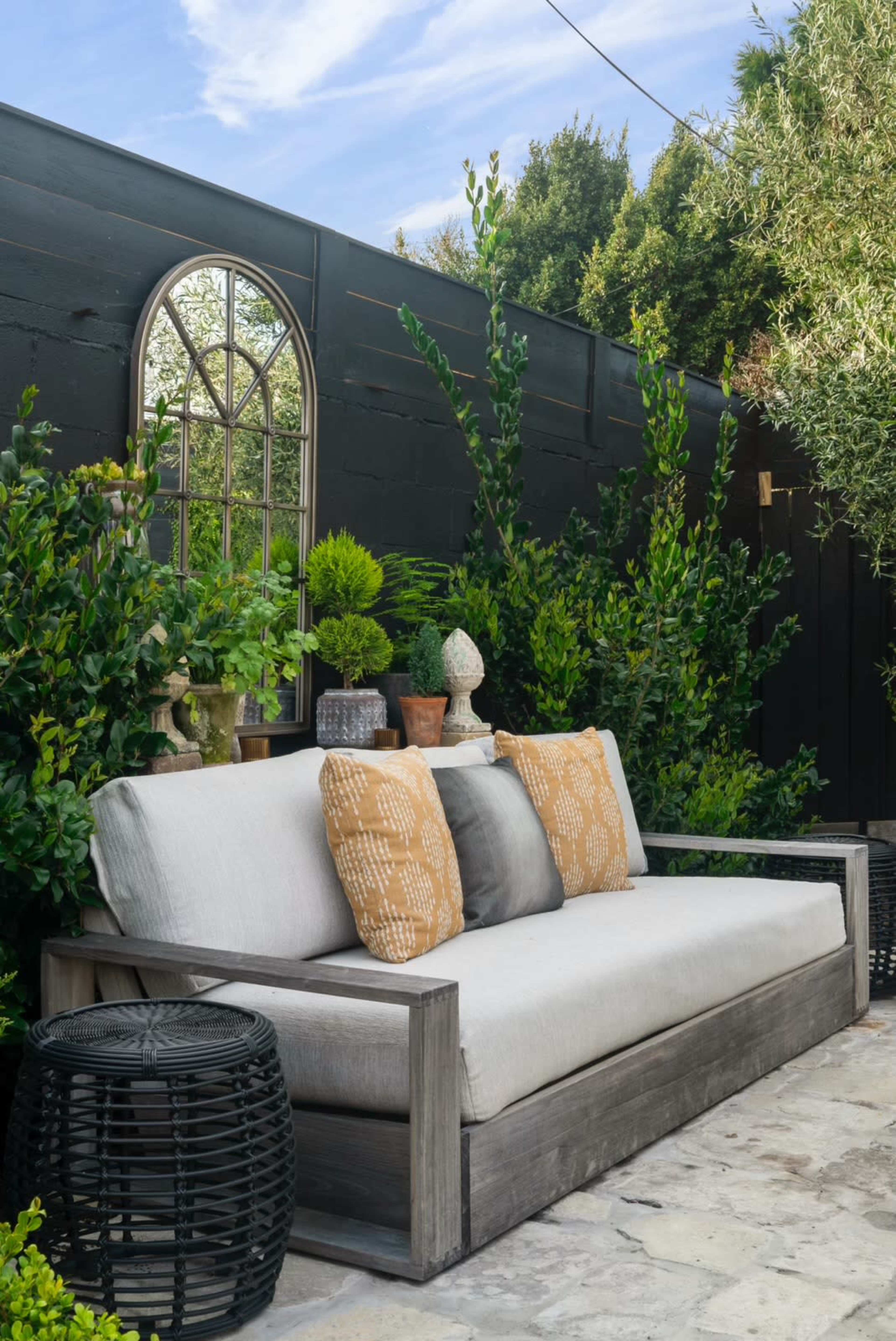 A light-colored outdoor sofa with decorative pillows is set against a dark wooden wall, surrounded by greenery and decorative plants.