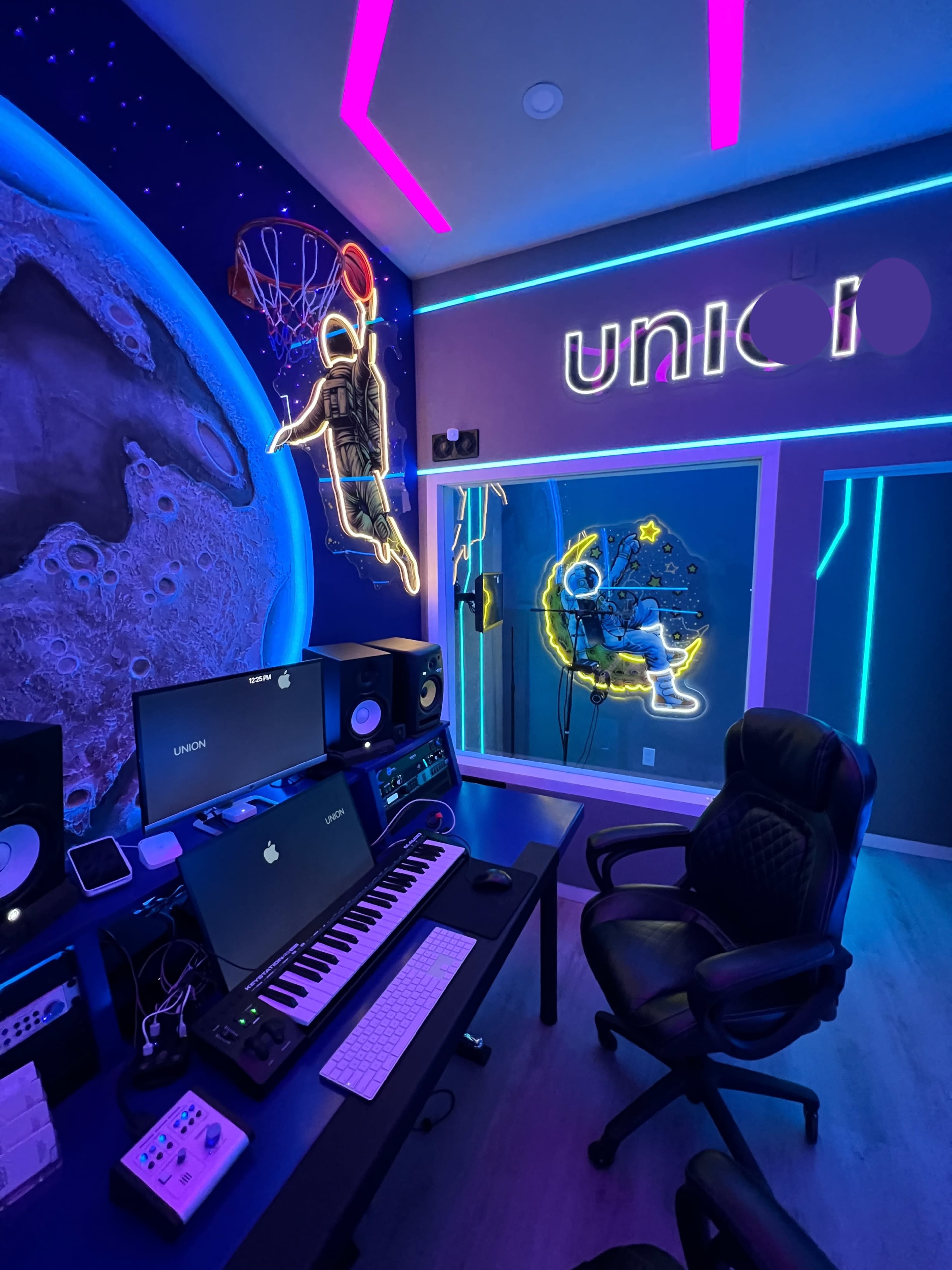 The image depicts a modern music studio featuring a neon-lit design with a desk, computer setup, and colorful wall art of an astronaut and a basketball player.
