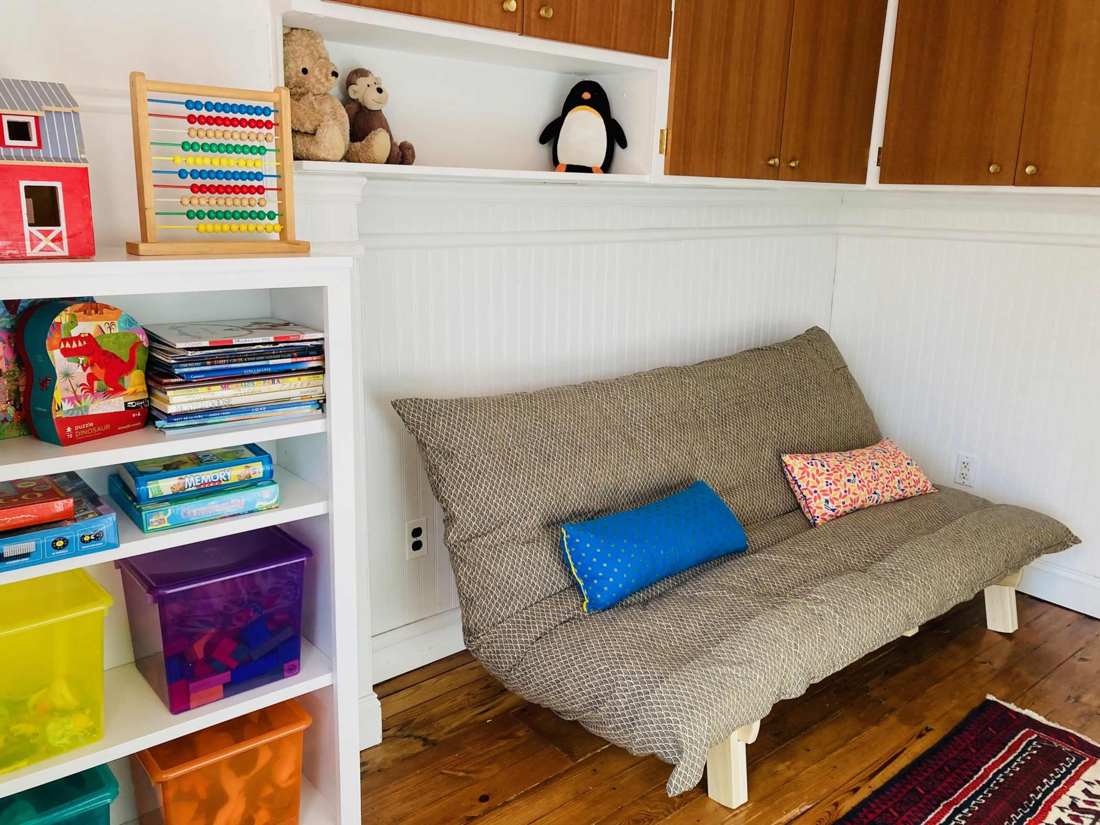 The image shows a children's play area featuring a small couch with decorative pillows, a shelf filled with toys and books, and colorful storage bins, all set against a bright wall.