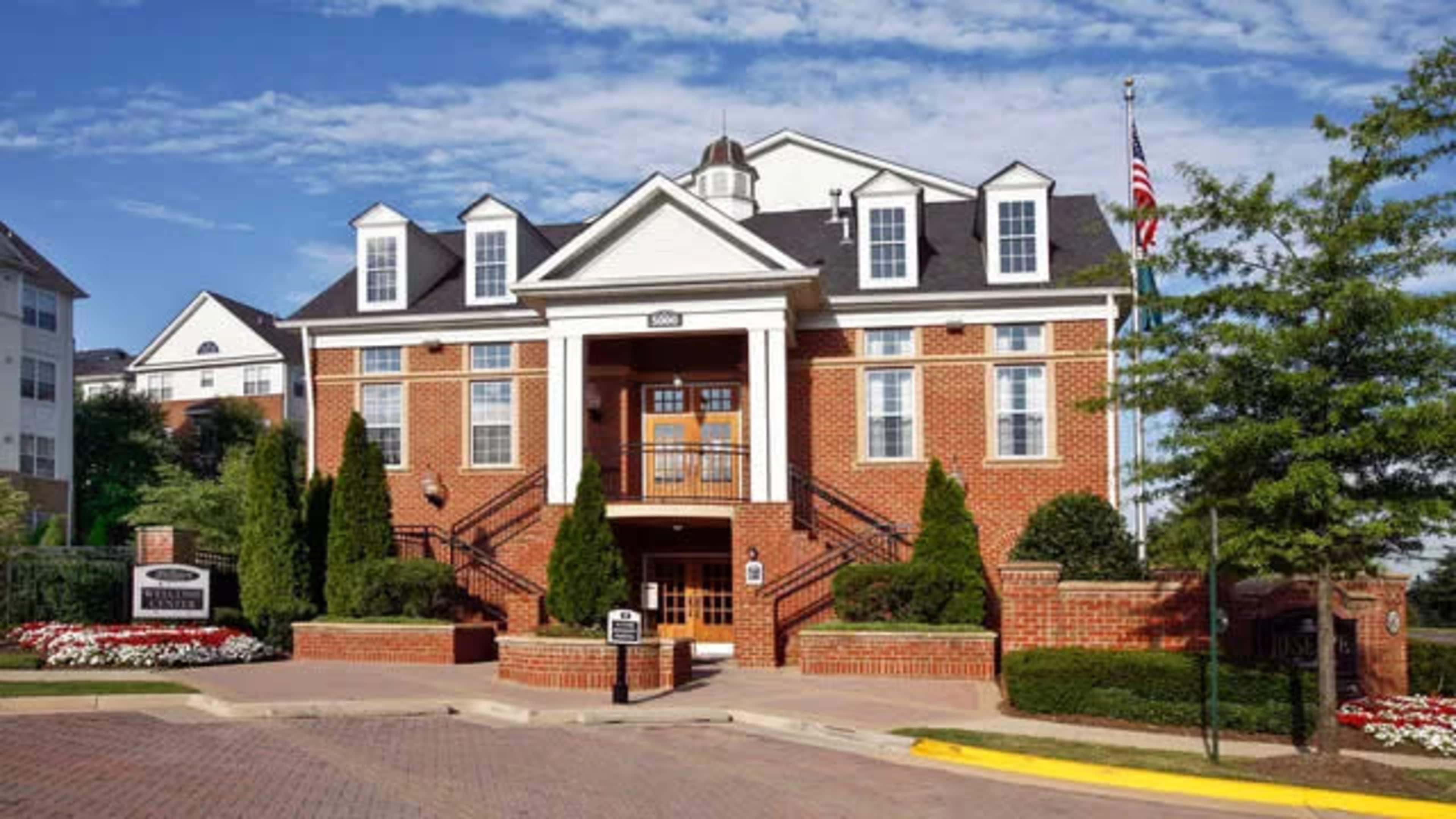 A brick building with a white-trimmed entrance, surrounded by neatly landscaped trees and flower beds.