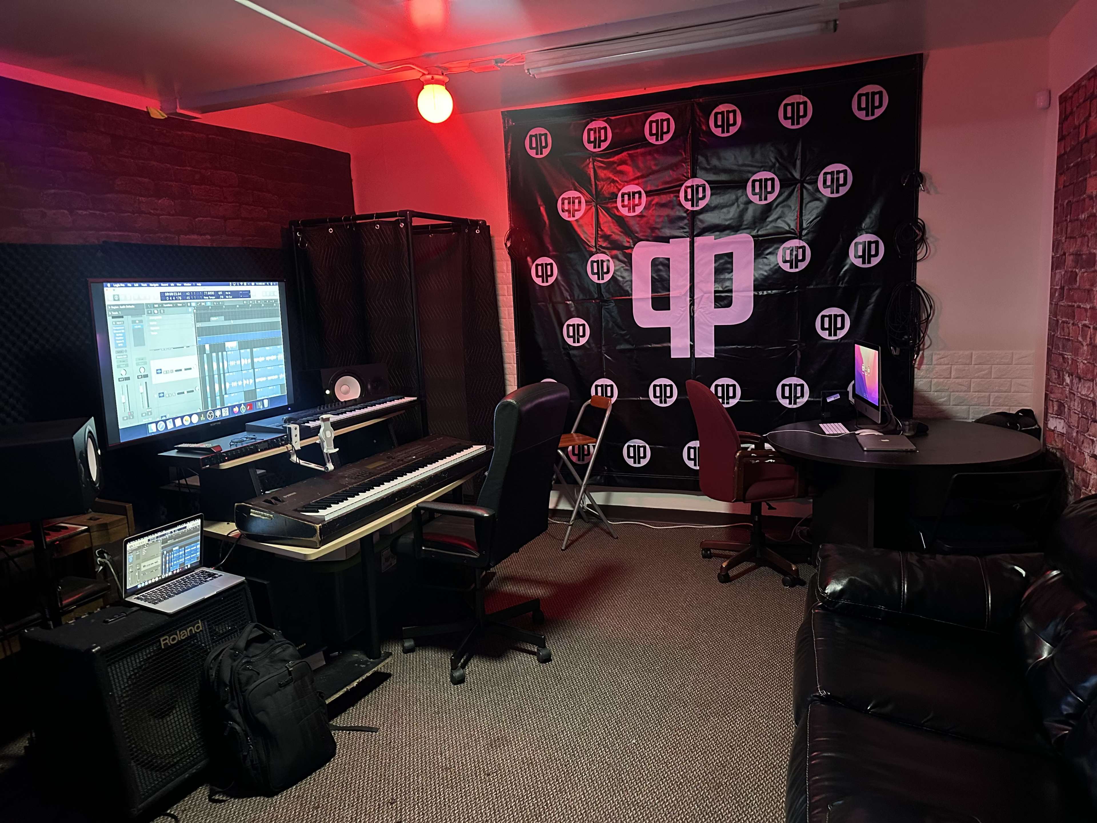 The image shows a music production studio with a keyboard, multiple screens, a large banner, and seating areas.