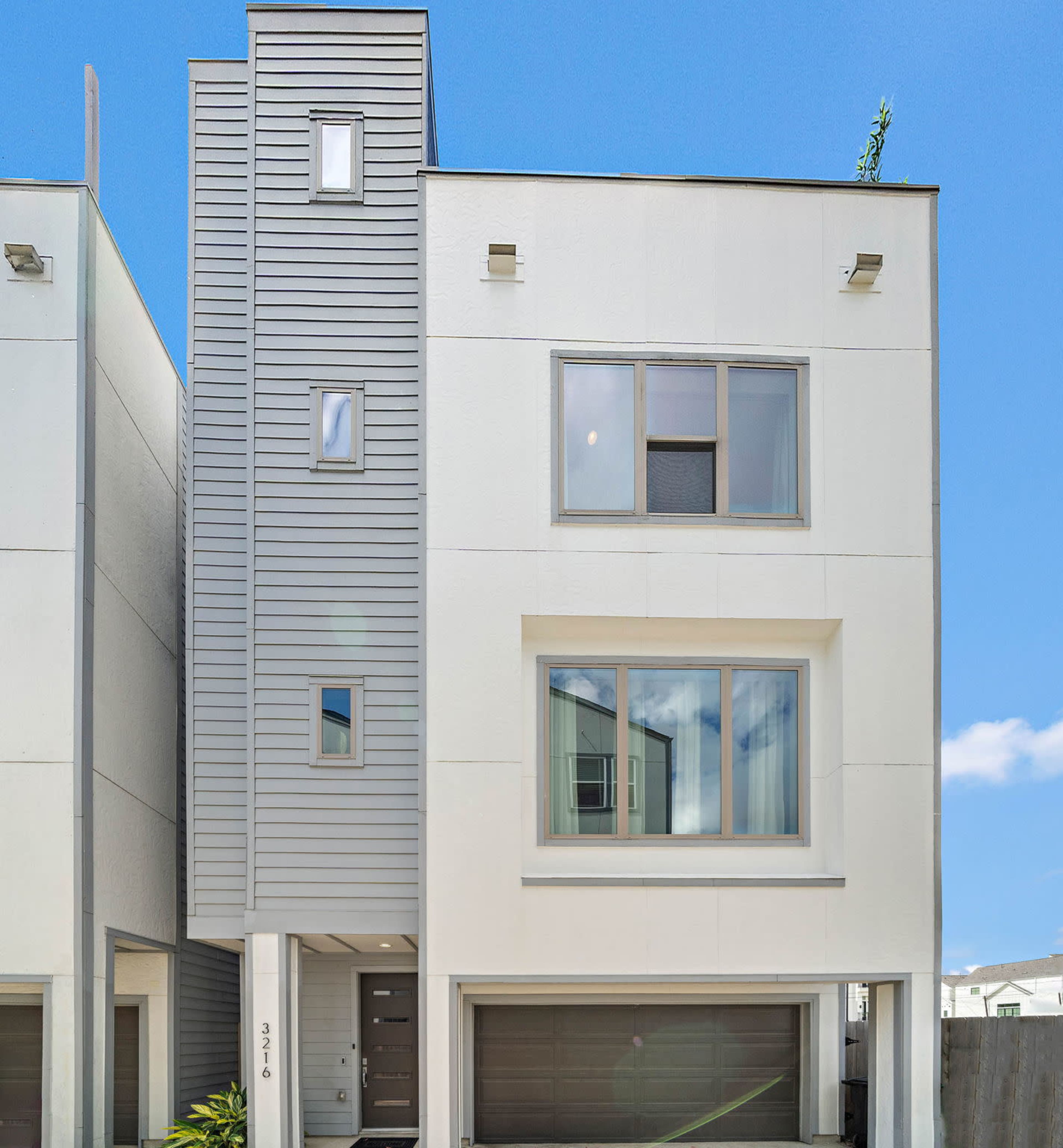 A modern three-story house with a mixed exterior of gray siding and white walls, featuring large windows and a garage door.