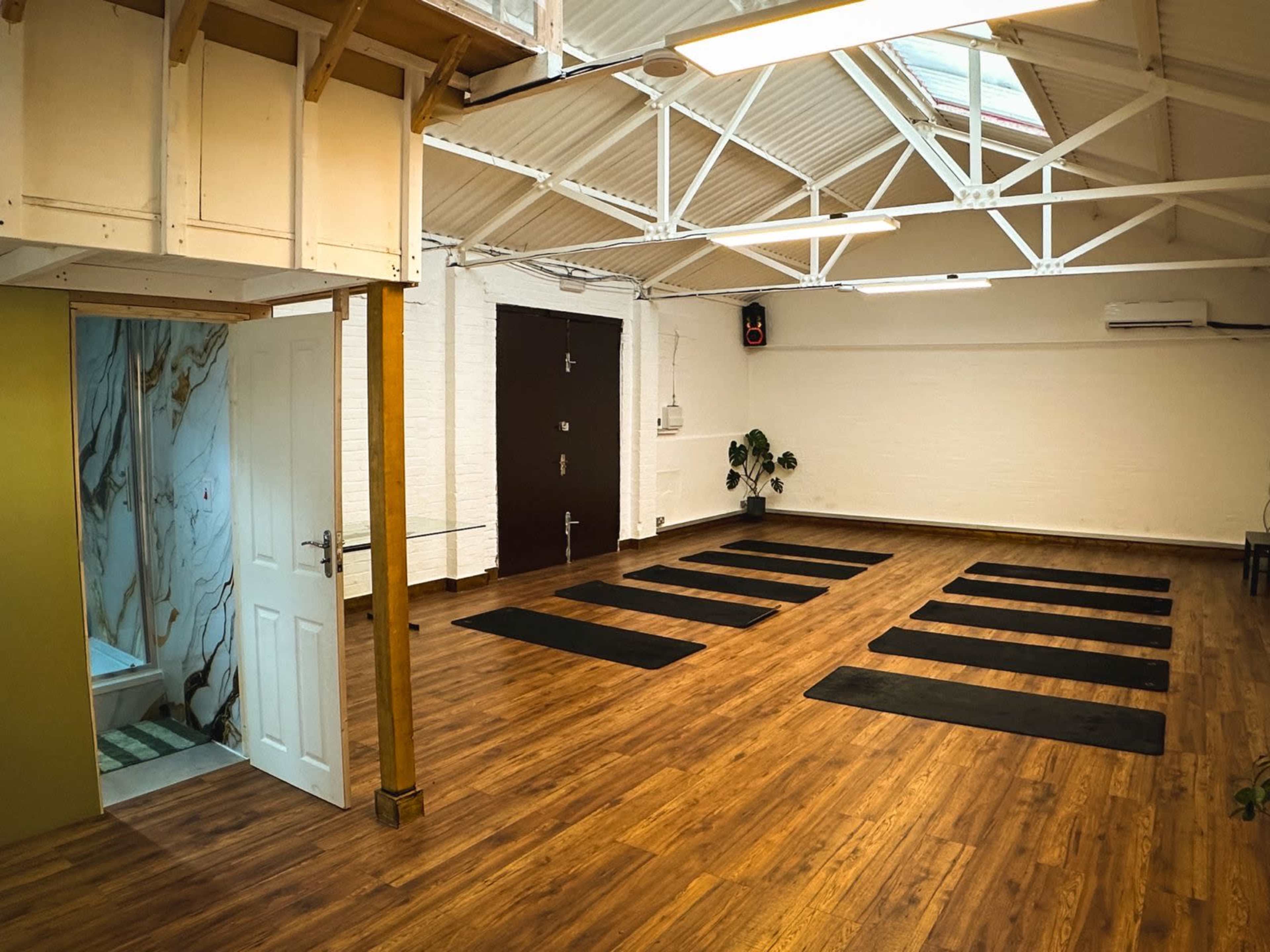 The image shows a spacious yoga studio with wooden flooring, several yoga mats arranged in parallel, and a small bathroom area visible to the left.