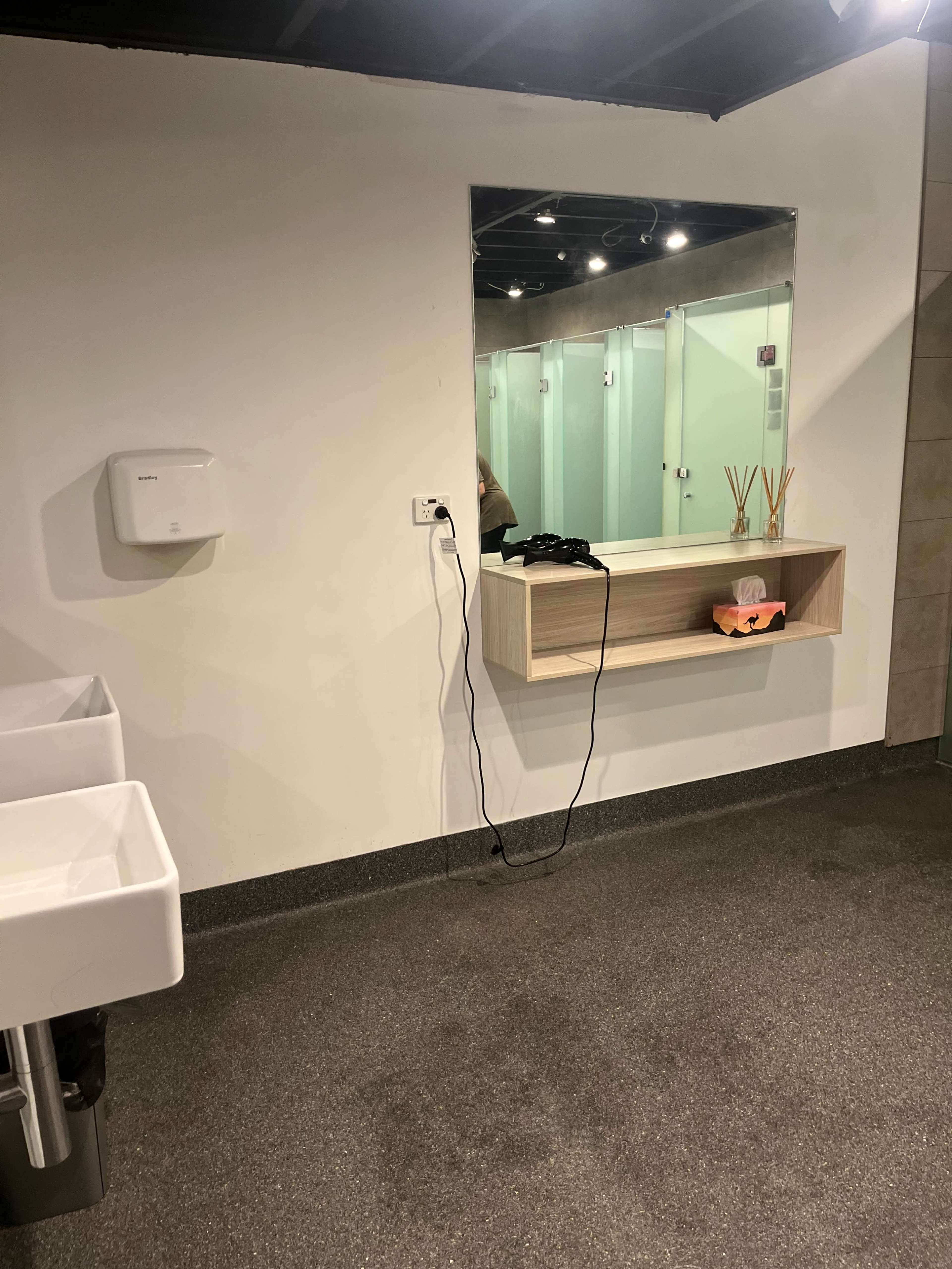 The image shows a bathroom area featuring two sinks, a large mirror, a shelf with decorative items, and several closed bathroom stalls in the background.