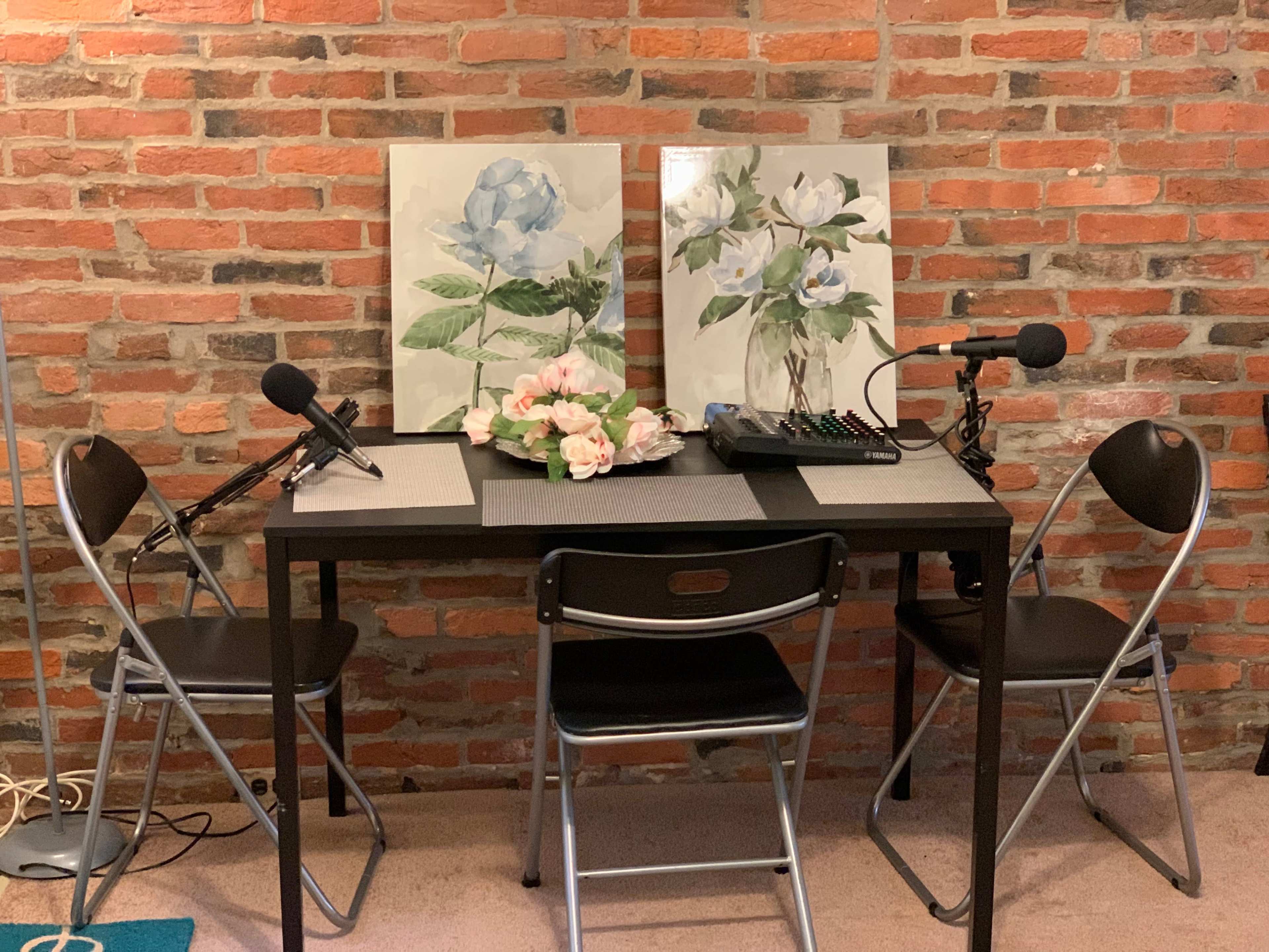 A small table with two framed floral artworks and two microphones is set up against a brick wall, with three black chairs arranged around it.