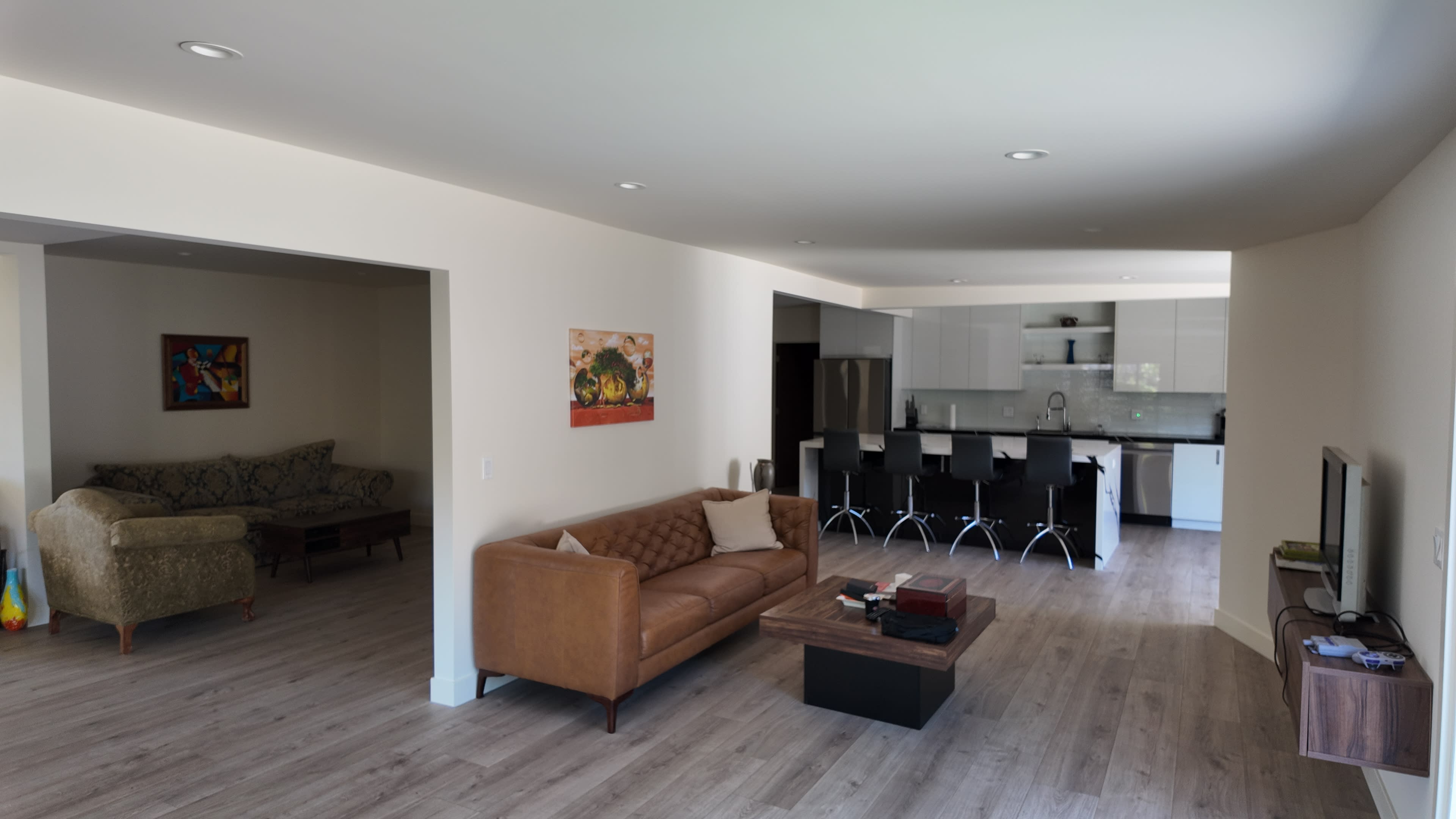 The image shows a modern living space featuring a brown leather sofa, a coffee table, and an open kitchen with bar stools in the background.