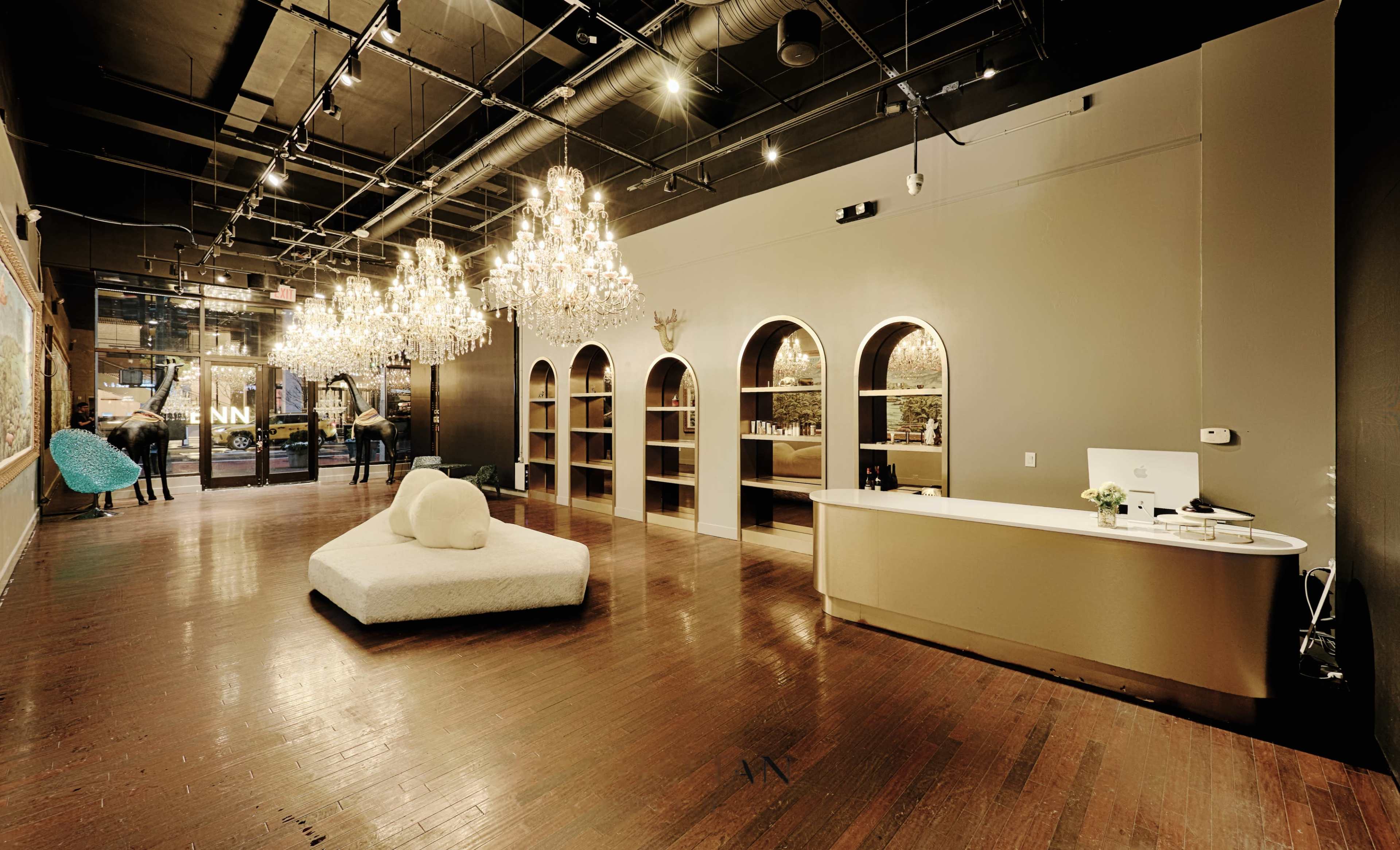 A spacious interior features a modern reception desk, vintage chandeliers, arched shelving units, and a large, plush seating area.