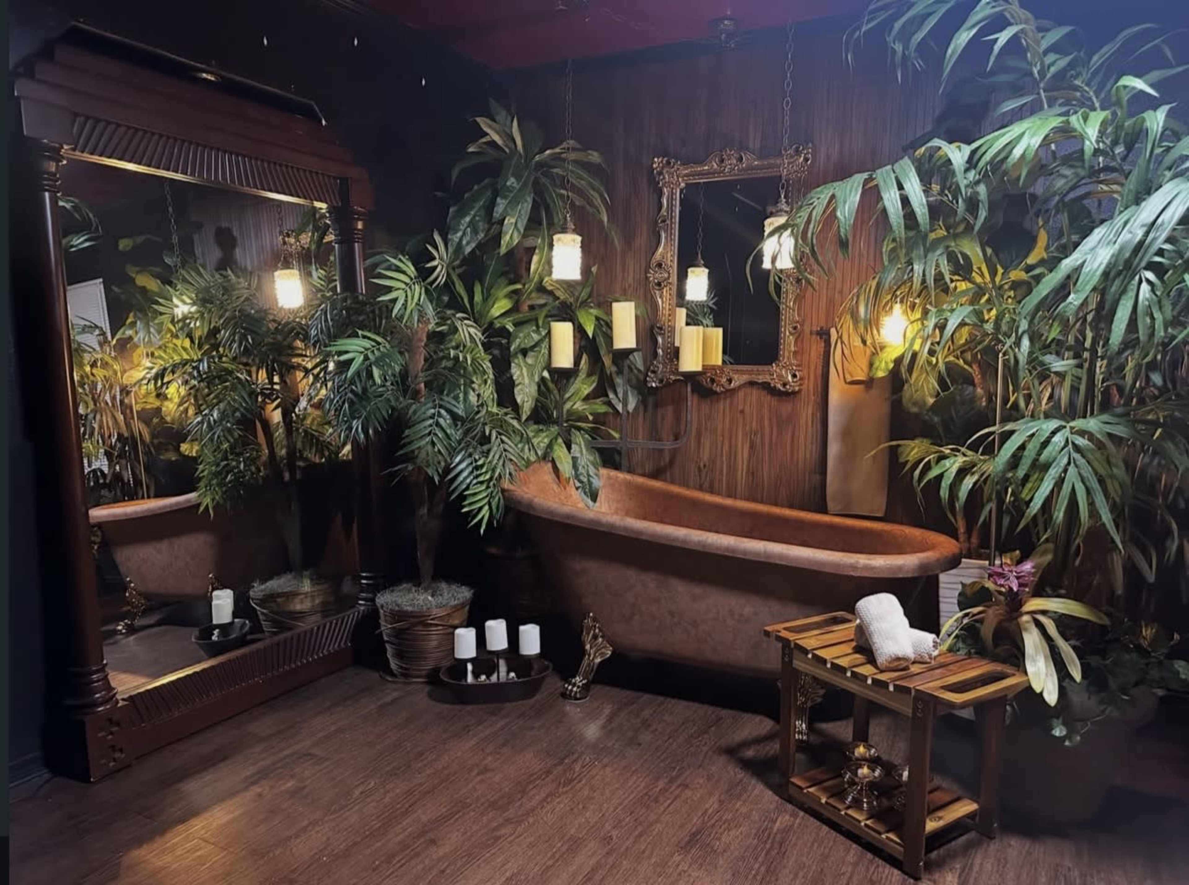 The image shows a cozy bathroom with a vintage bathtub, a large mirror, lush plants, and warm lighting.