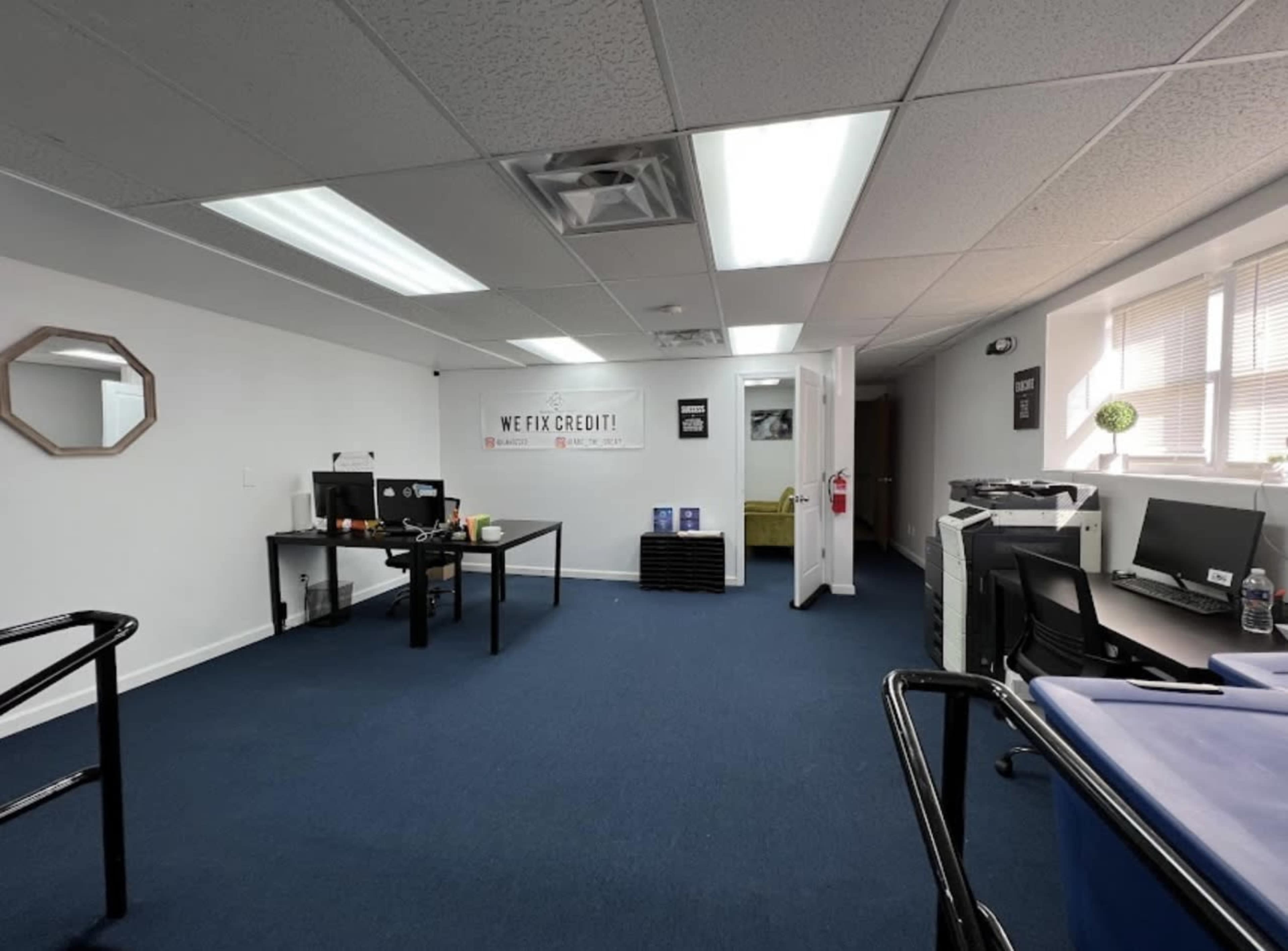 A tidy office space with two desks, computers, a blue carpet, and a wall featuring a motivational sign that reads "WE FIX CREDIT!"