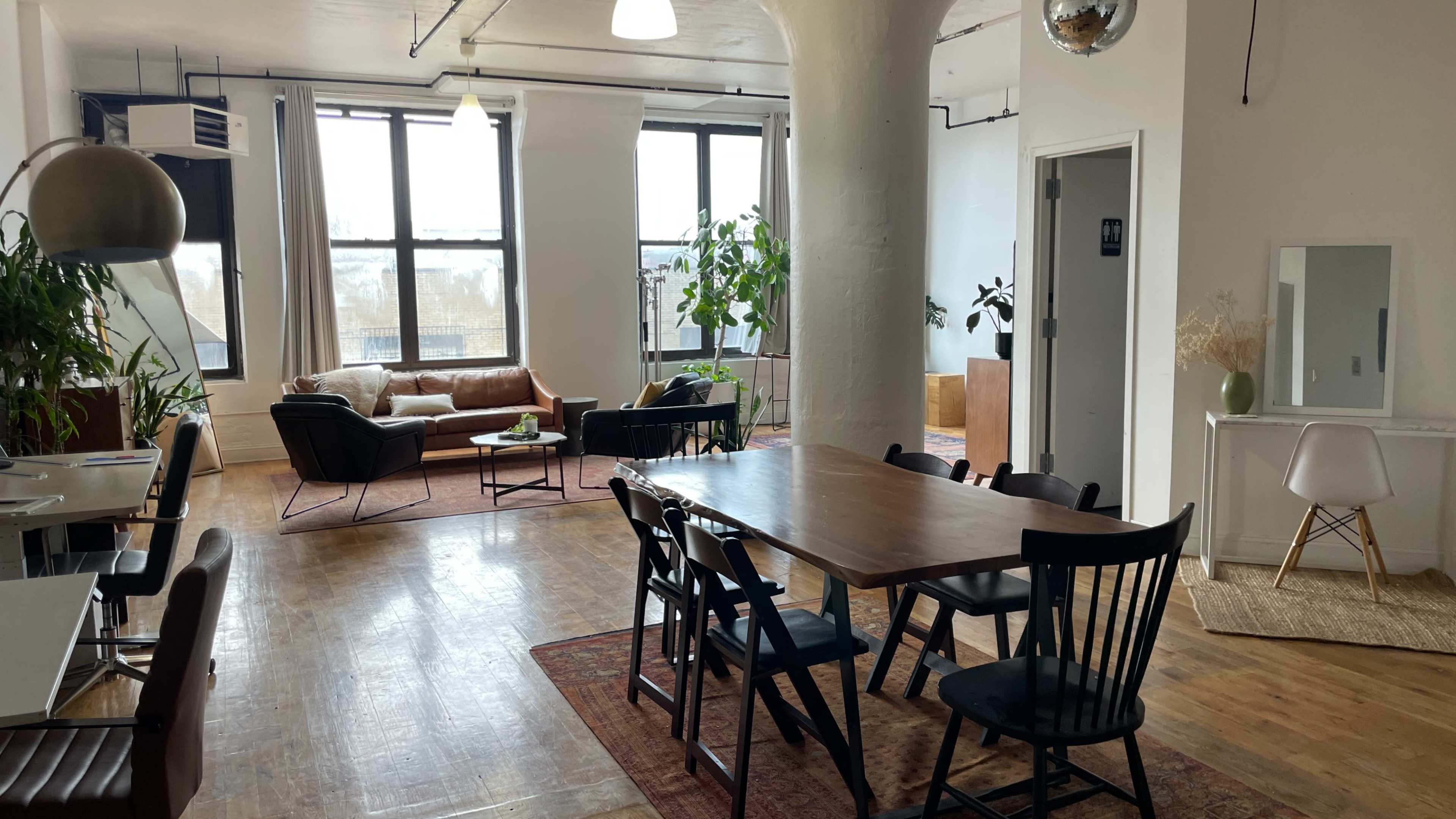 The image shows a spacious, modern room with a dining table and chairs, a living area featuring a brown leather sofa, and large windows allowing natural light to enter.