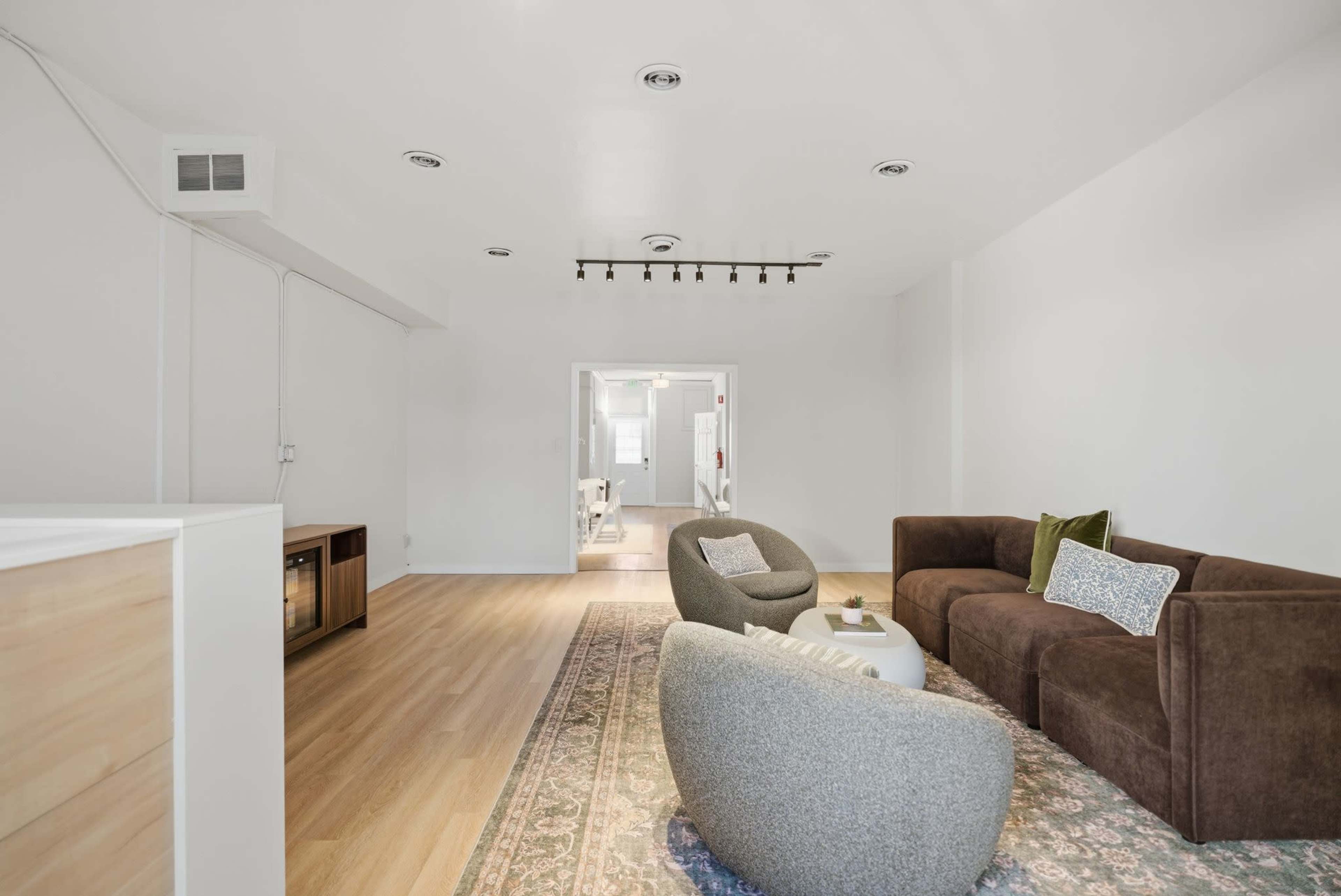 The image shows a modern living room with light wooden flooring, two distinct seating areas featuring a brown sectional sofa and gray armchairs, and a subtle area rug.