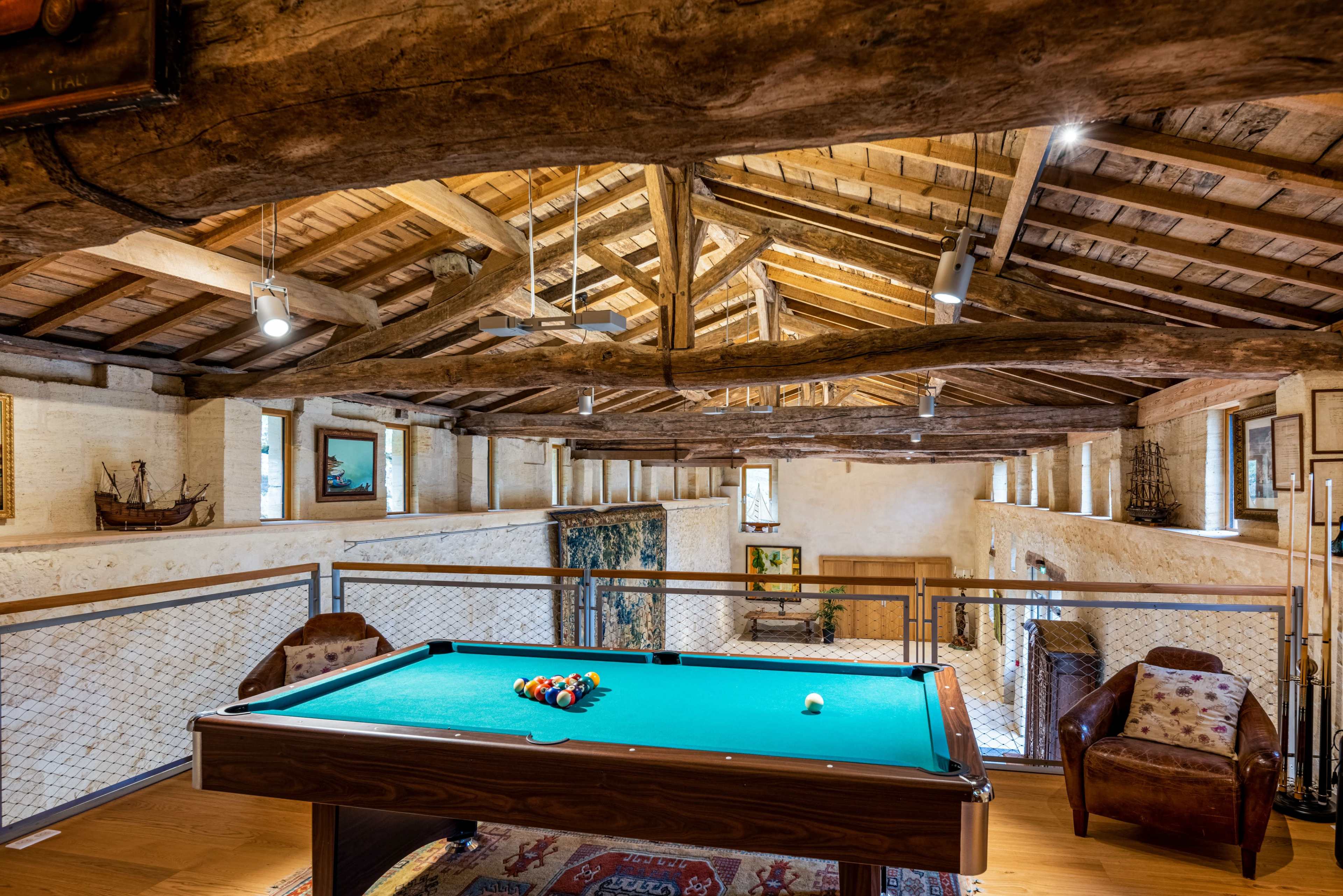 The image shows a spacious room with wooden beams and a green pool table in the center, surrounded by rustic decor and framed artwork.