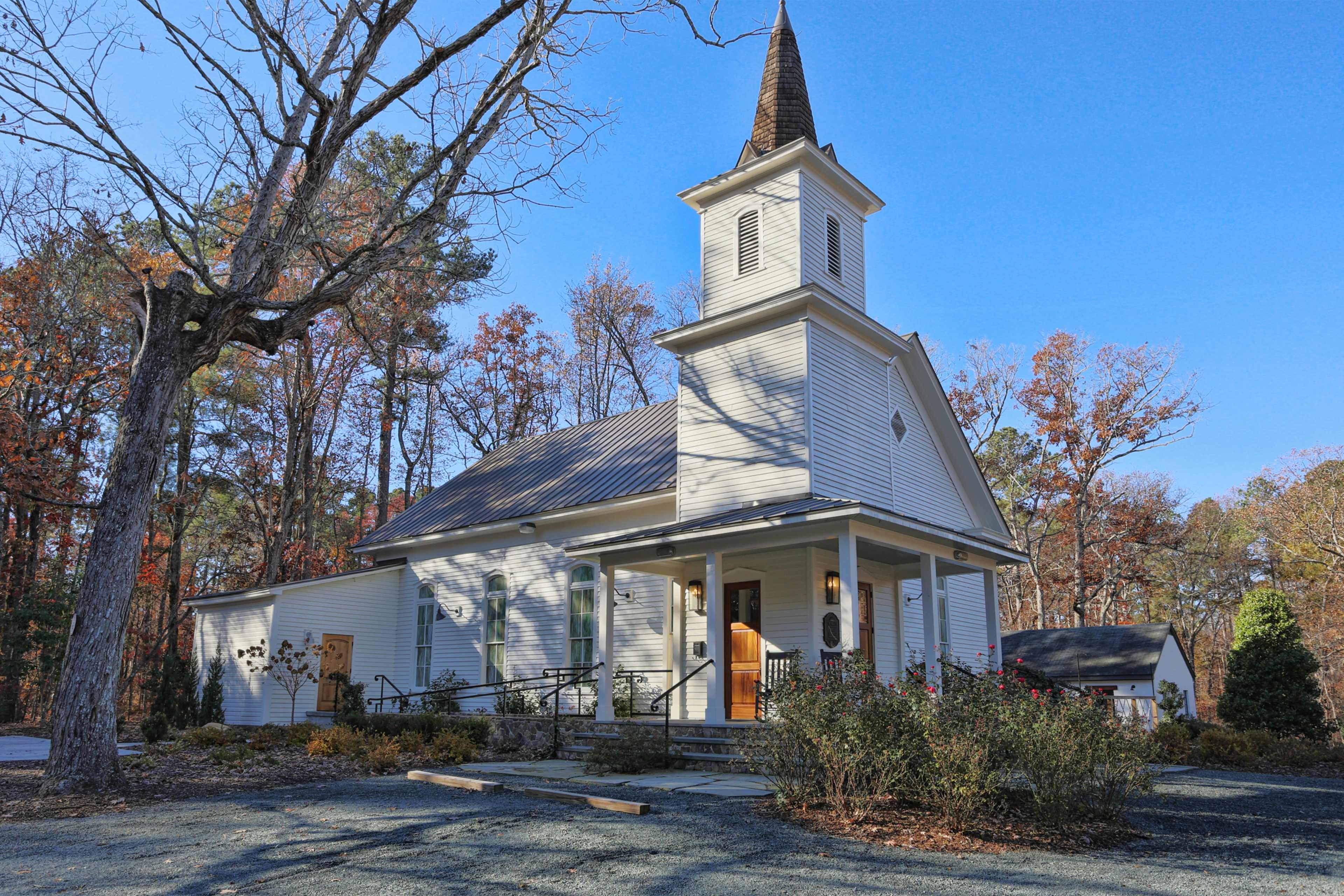 A white church with a tall spire, surrounded by trees and a gravel walkway.