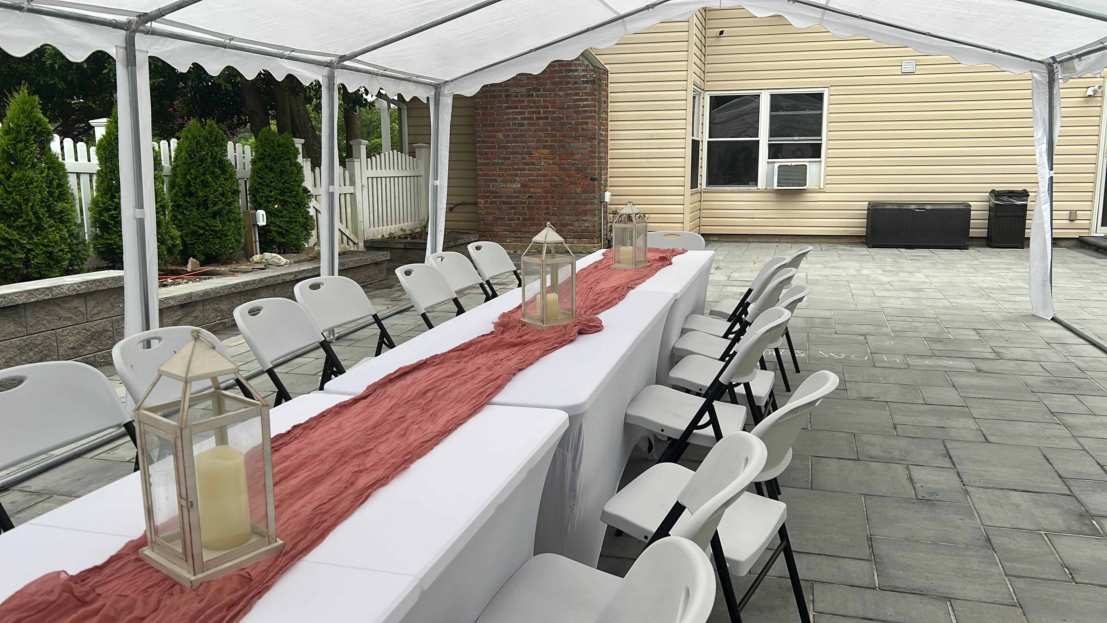A long table is set up under a white tent, flanked by folding chairs and adorned with lanterns along a pink runner, with a house and greenery in the background.