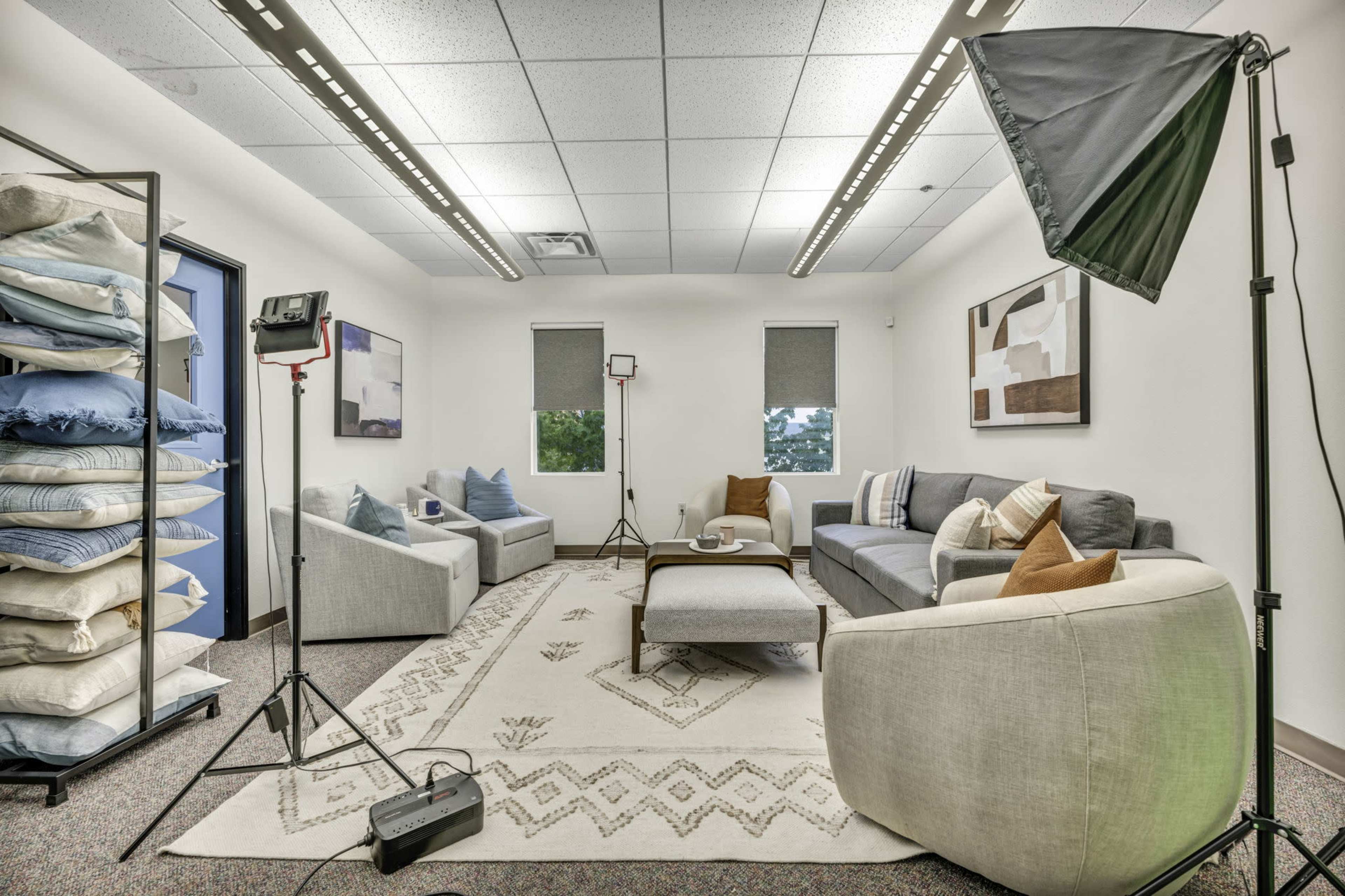 A well-lit room features two couches facing each other, a coffee table in the center, and a stack of pillows against one wall, with lighting equipment set up for a photo shoot.