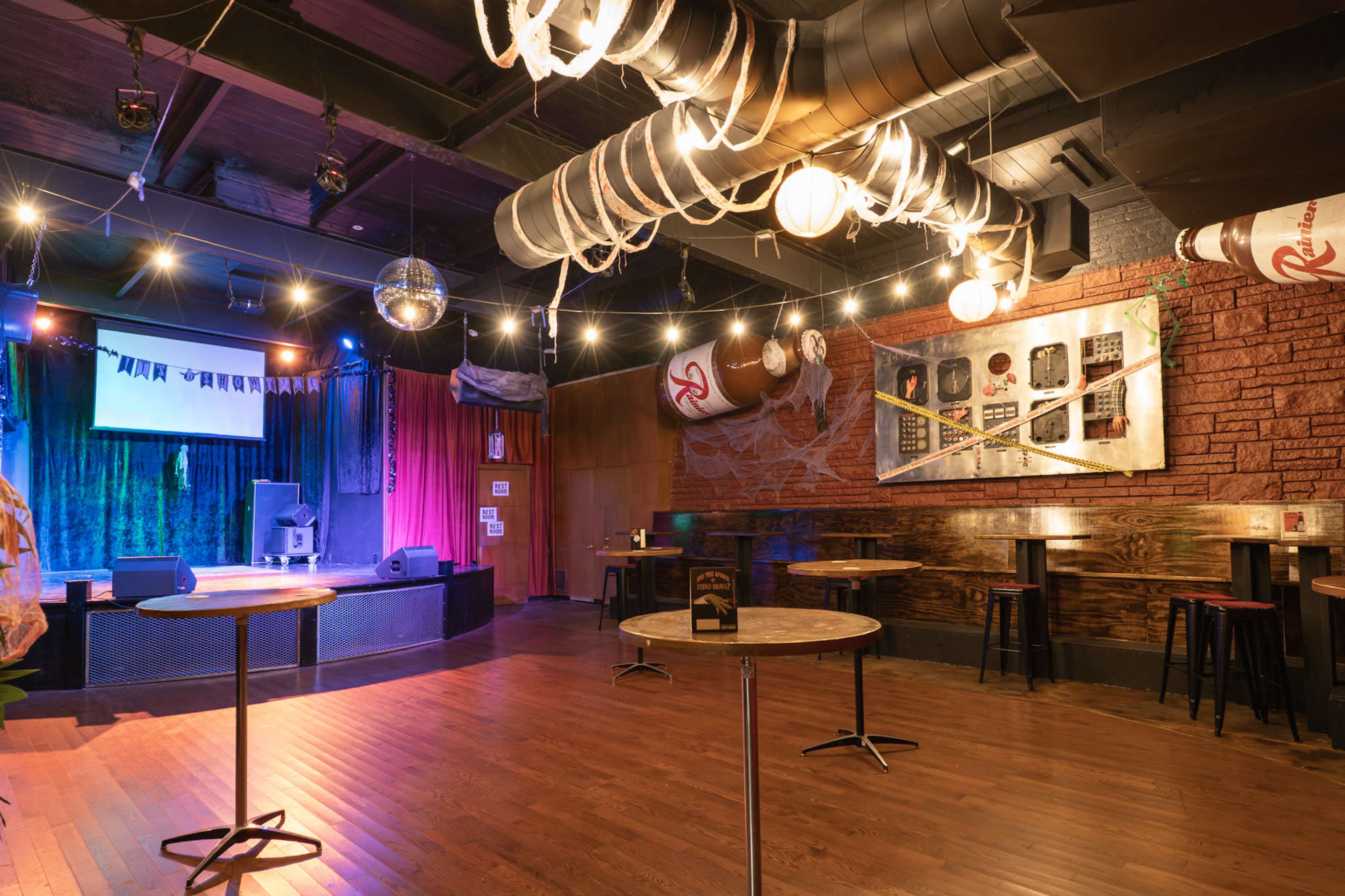 The image shows an empty performance venue with a raised stage, dim lighting, and scattered tables.