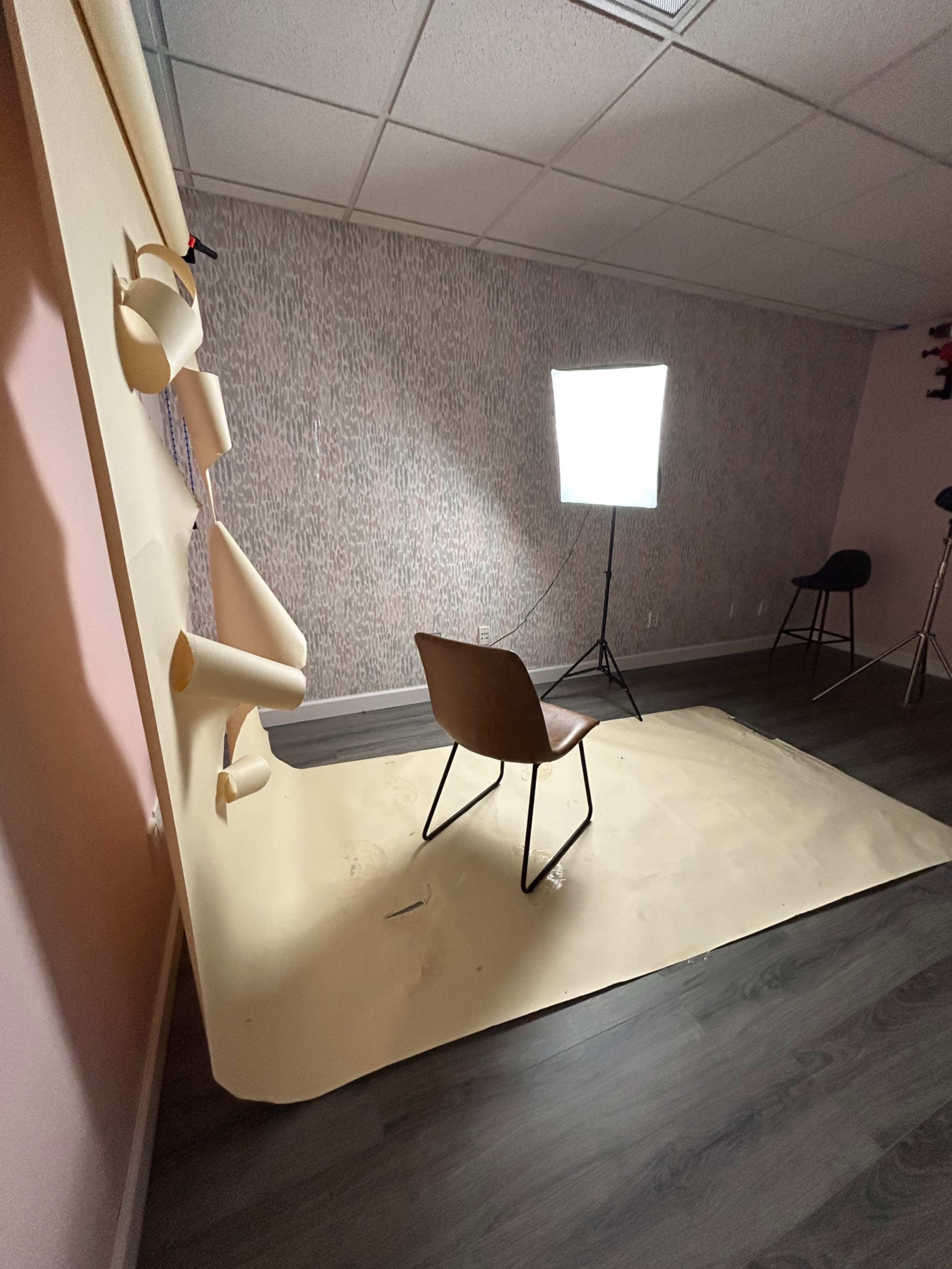 The image shows a sparsely furnished photography studio with a chair positioned on a large sheet of beige paper, illuminated by a softbox light against a patterned wall.
