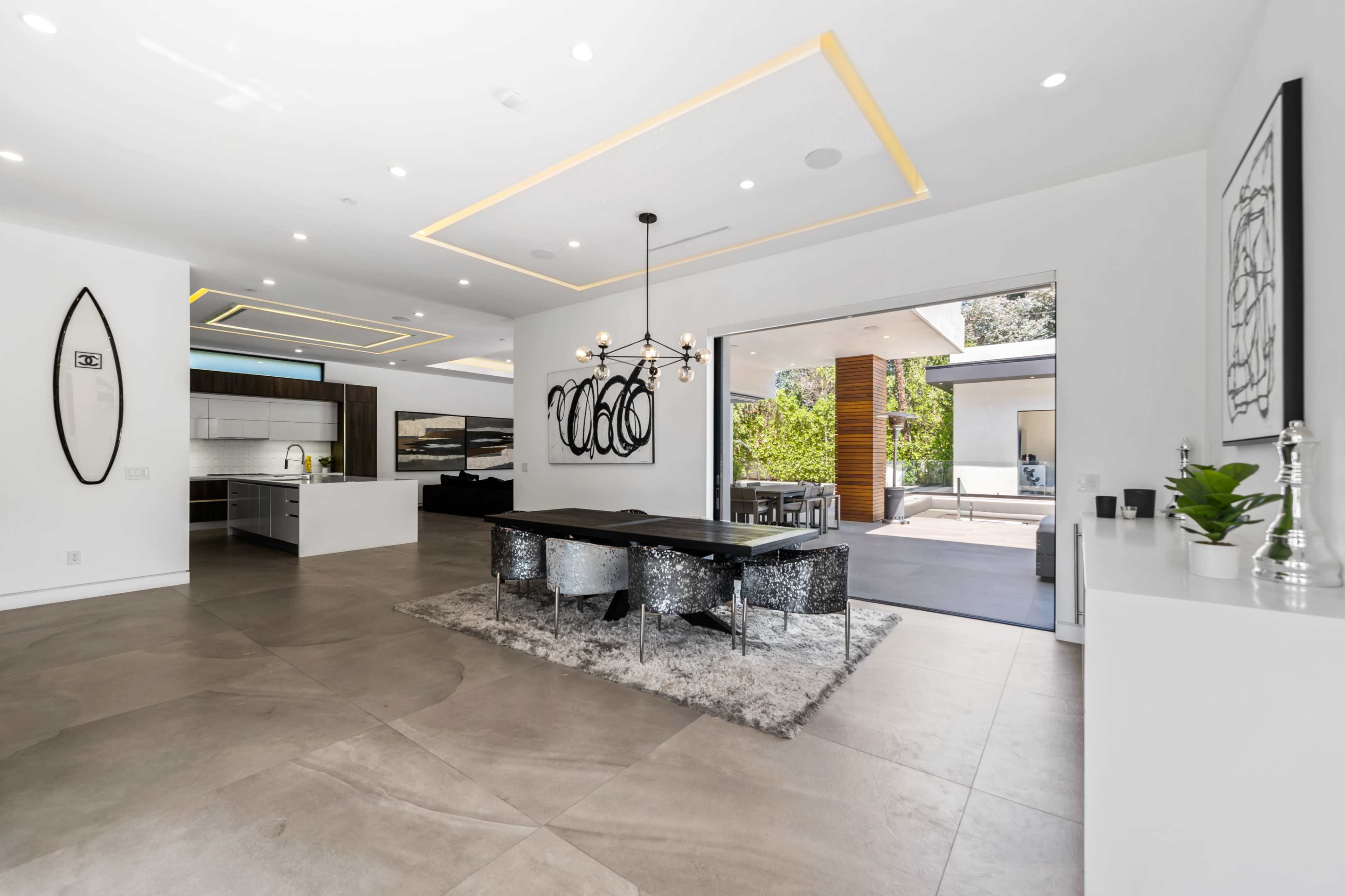 A modern open-concept living space features a dining area with a sleek table and chairs, illuminated by a geometric chandelier, leading to an outdoor patio.