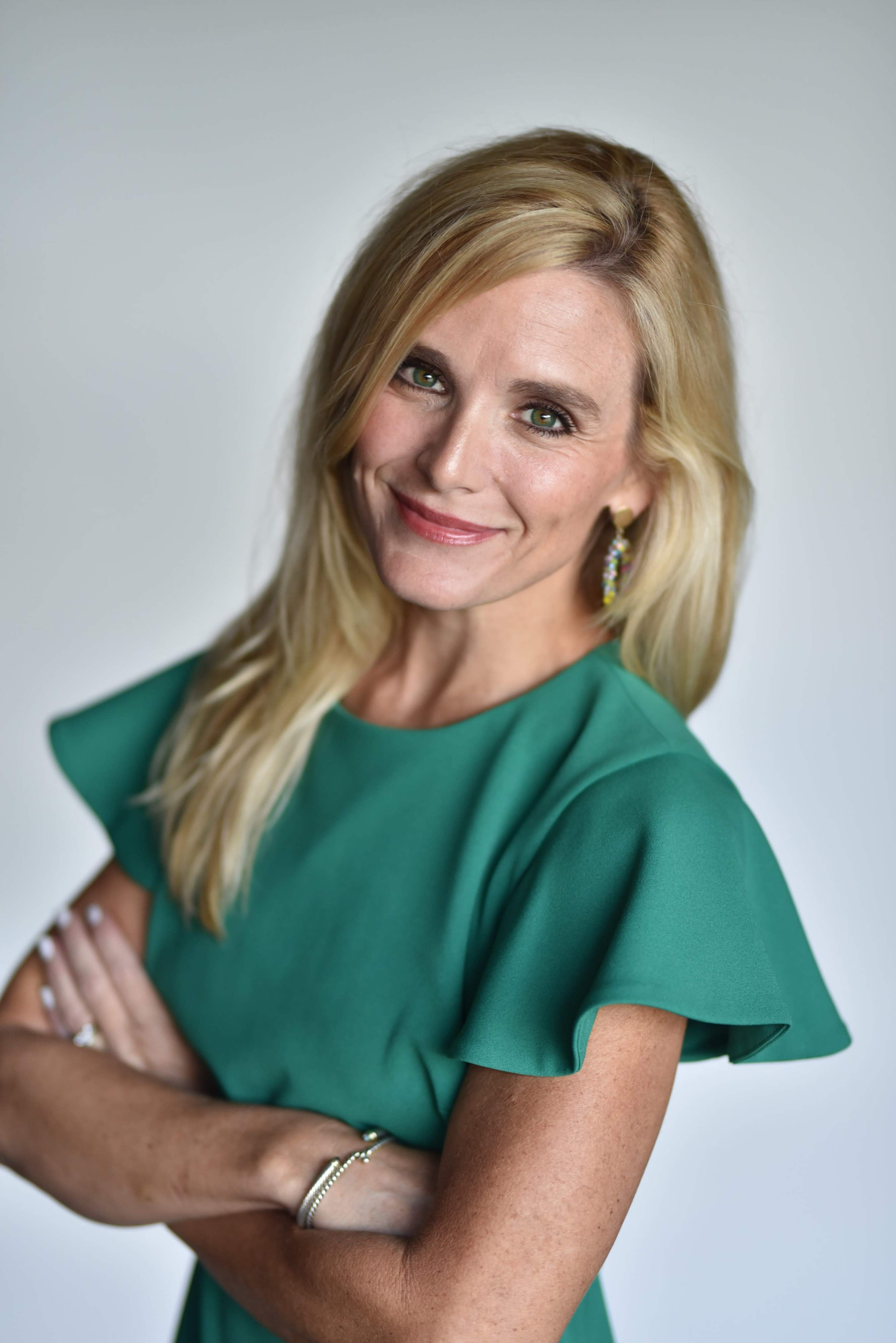 A woman with long blonde hair stands confidently, wearing a green dress with ruffled sleeves and smiling at the camera.