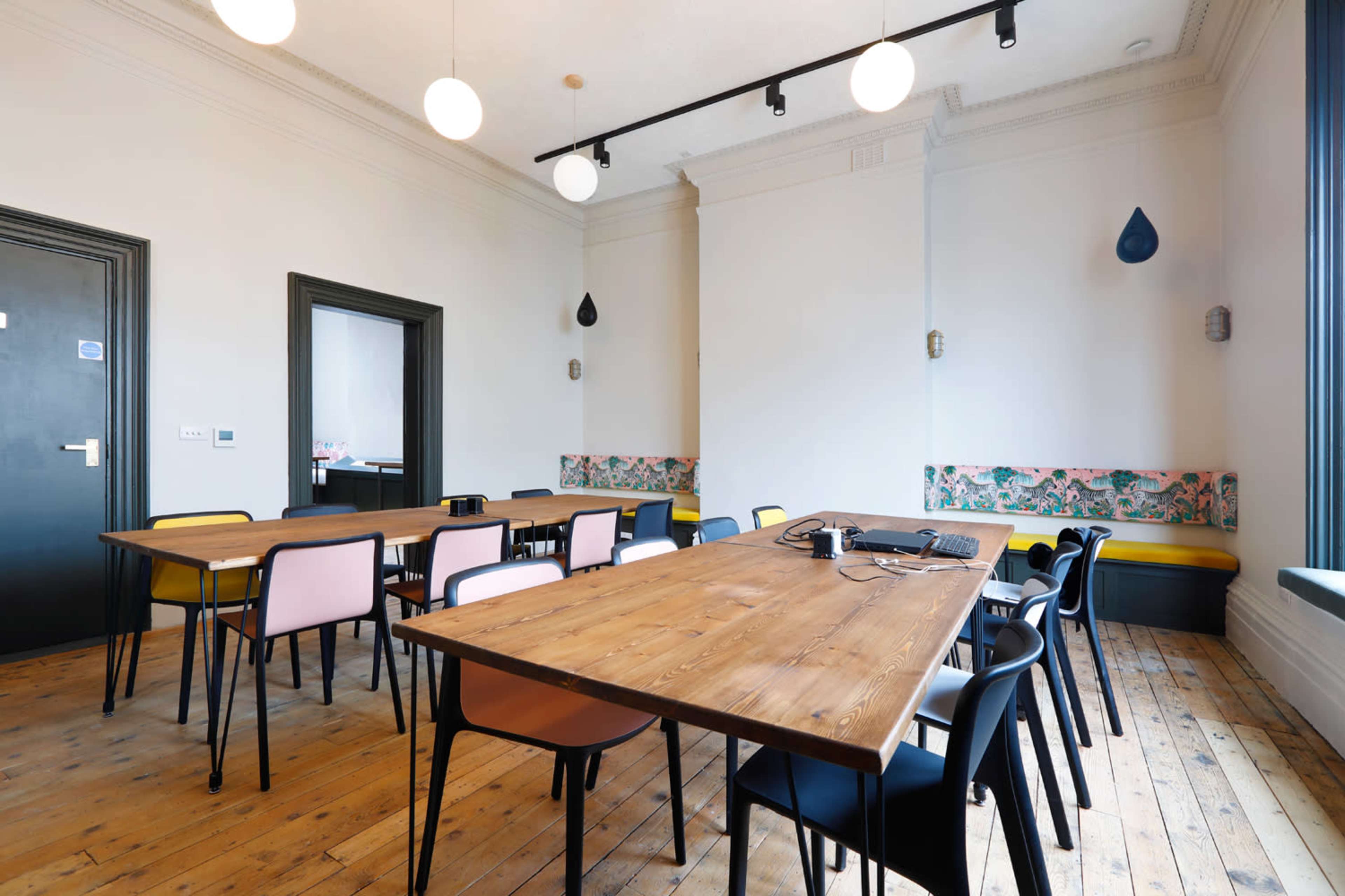 A spacious meeting room features several wooden tables surrounded by colorful chairs, with wall decor and pendant lighting.