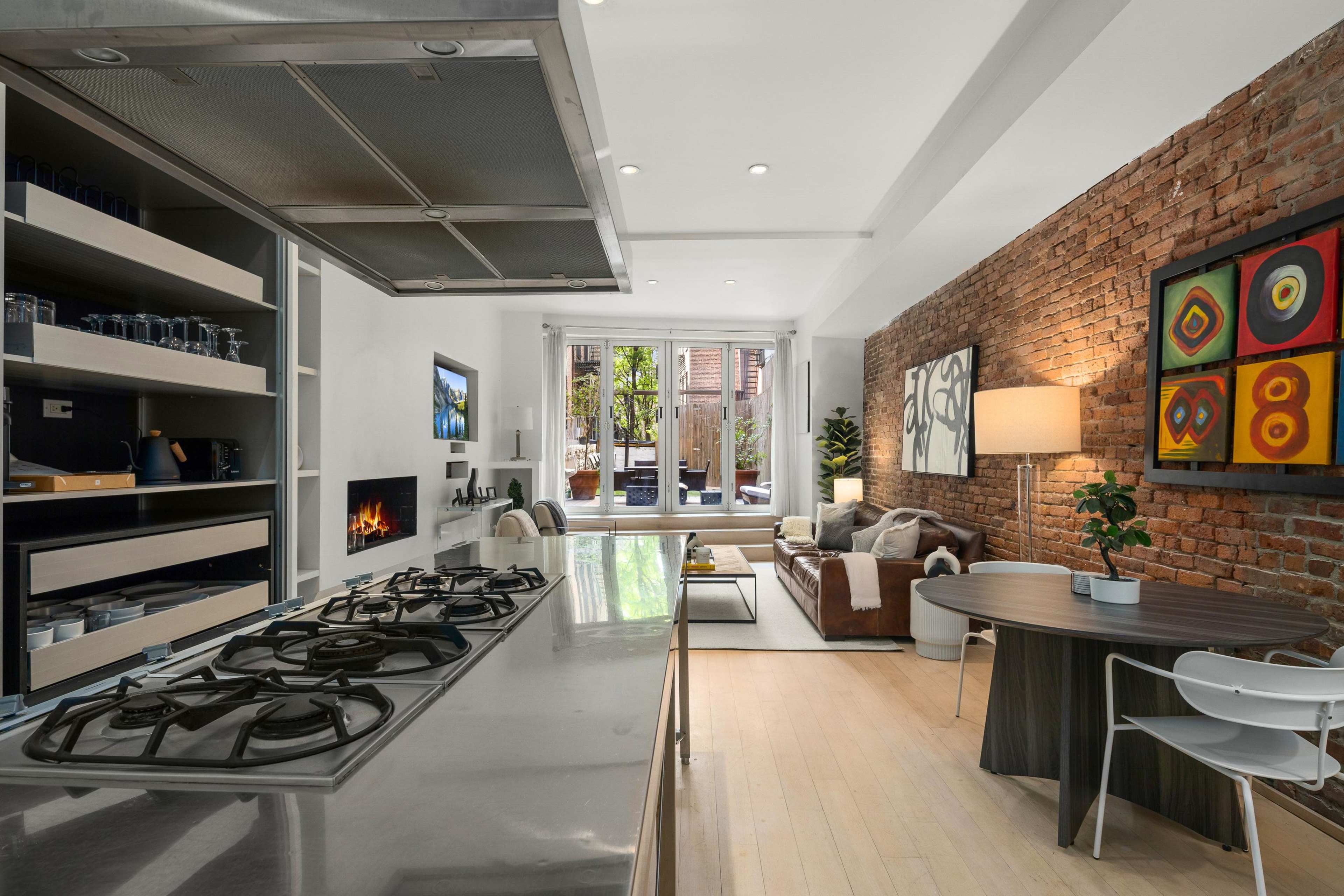 A modern kitchen with a stainless-steel countertop and gas stove, adjacent to a living area featuring a brown sofa and a view of an outdoor space through large windows.