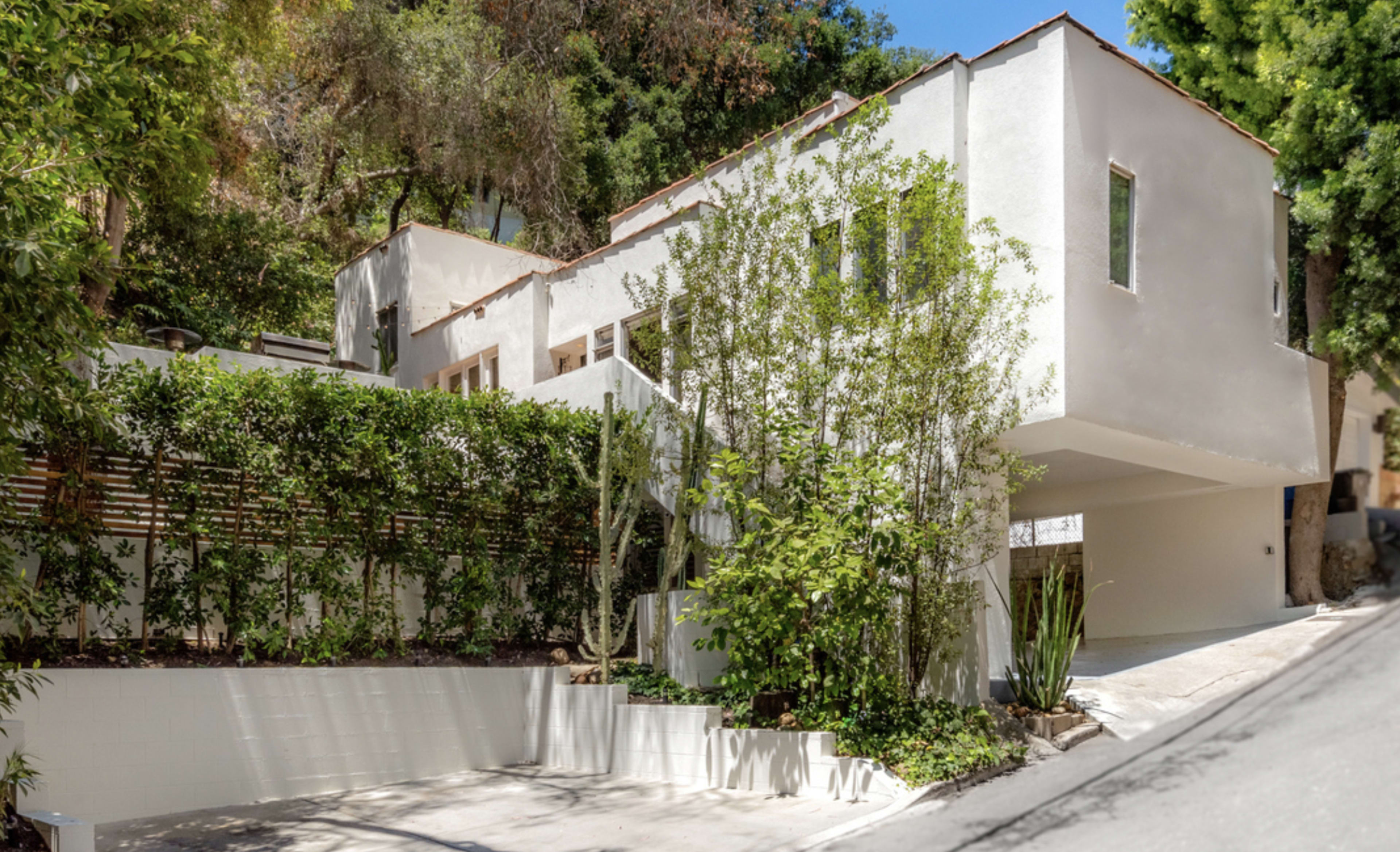 A modern white house is set on a sloped lot, surrounded by greenery and featuring a driveway at the front.