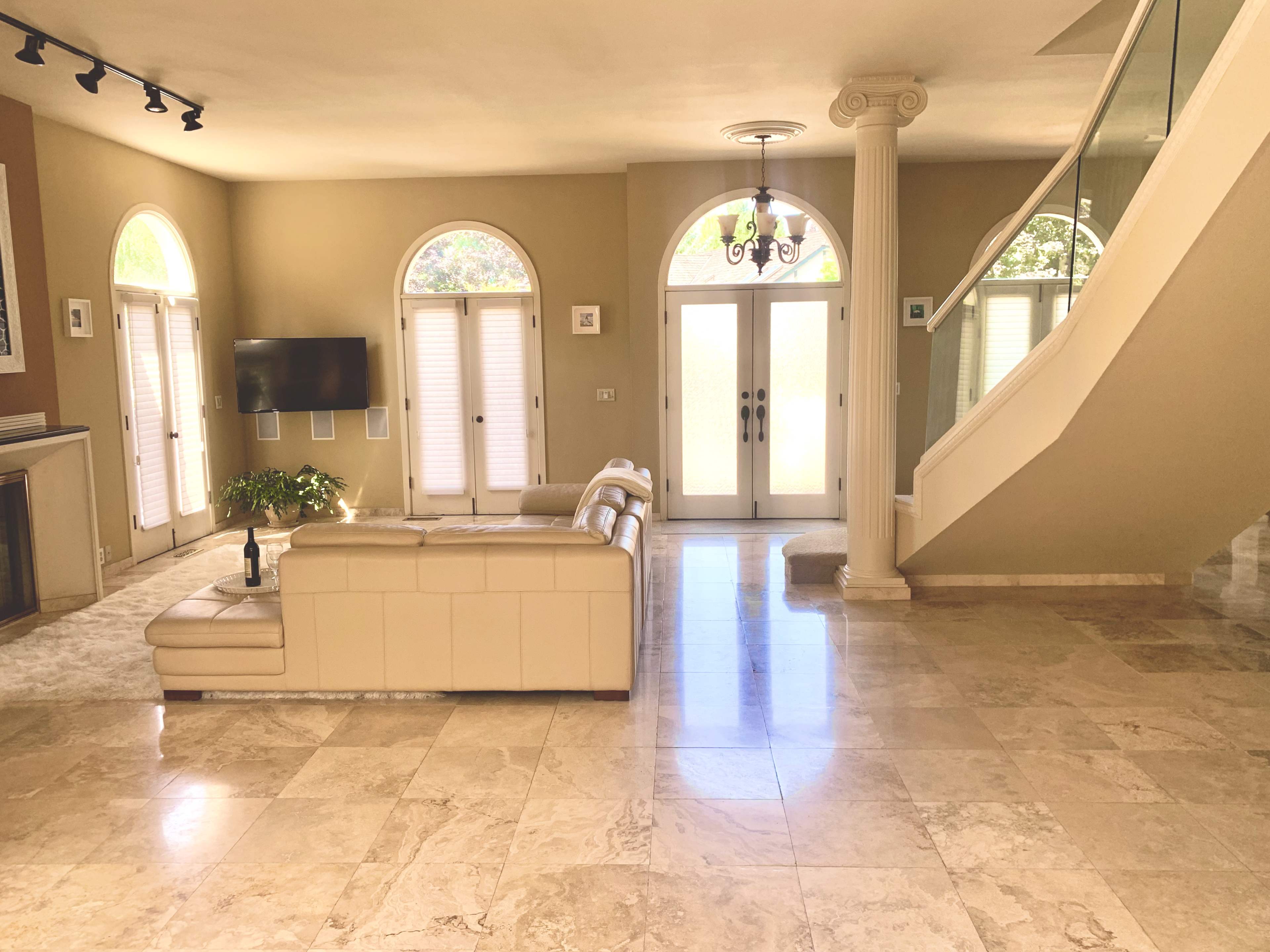 A spacious living area with a cream-colored sofa, a television mounted on the wall, large windows flanked by shutters, and a staircase leading to an upper level.