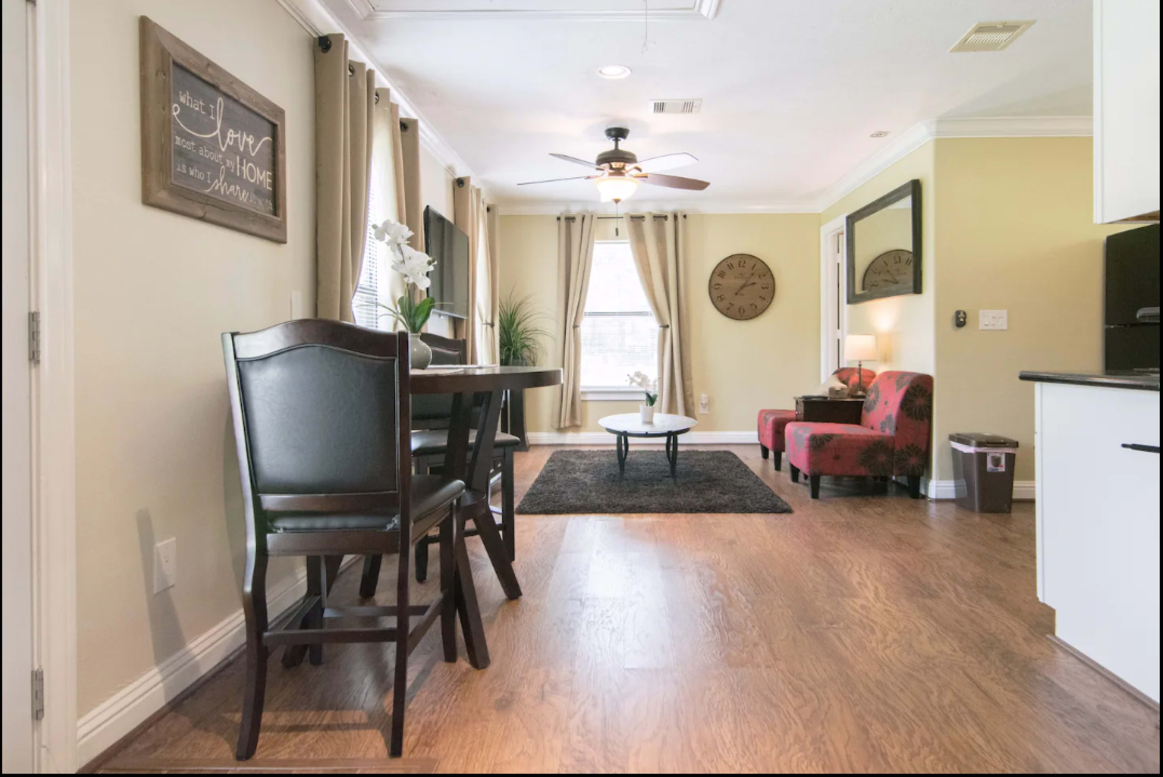 A living area with a dining table and chairs, a coffee table on a rug, and two red chairs, all illuminated by natural light from a window.