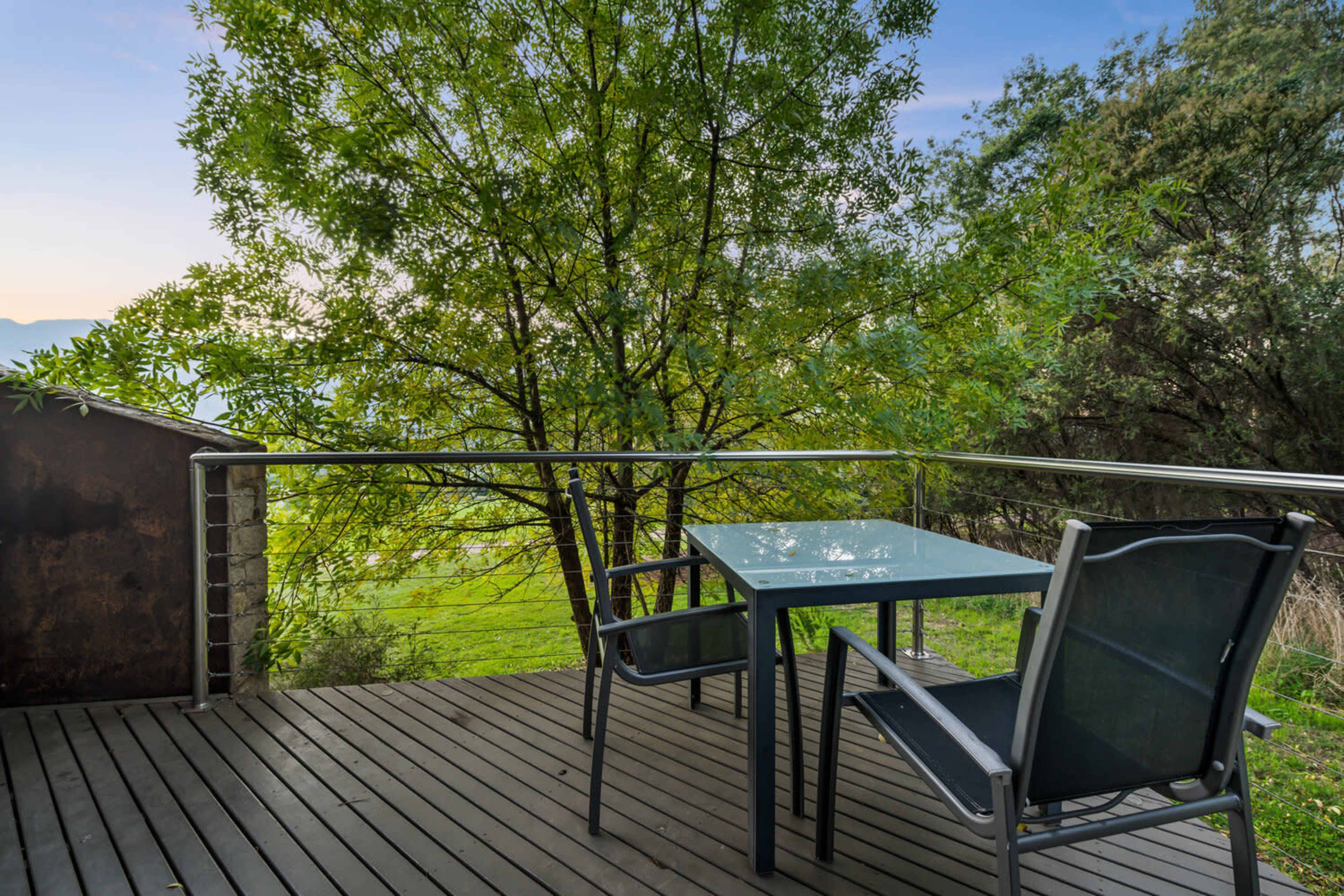 A metal table and two chairs are positioned on a wooden deck surrounded by green foliage and an expansive view of grass and hills.