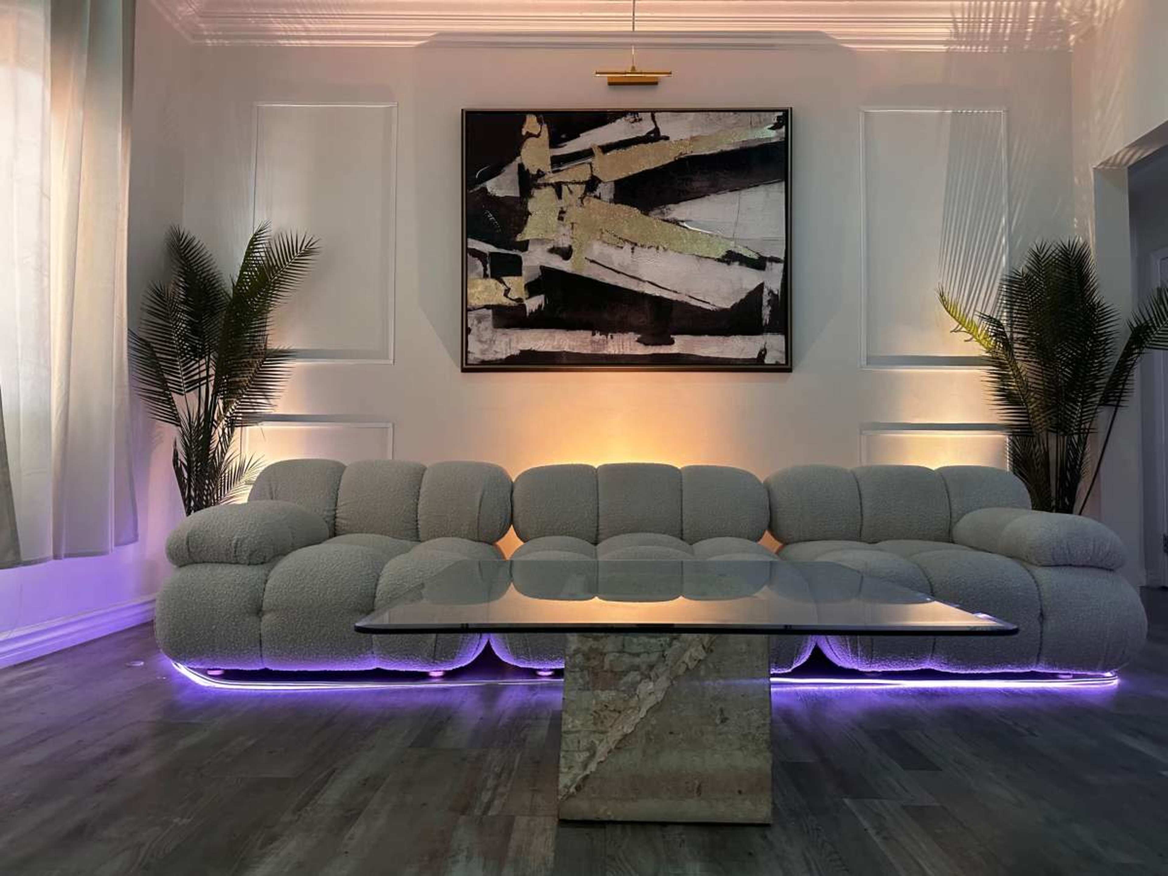 A modern living room features a plush, light-colored sectional sofa with glowing edges, a glass-top coffee table, and a large abstract painting on the wall.