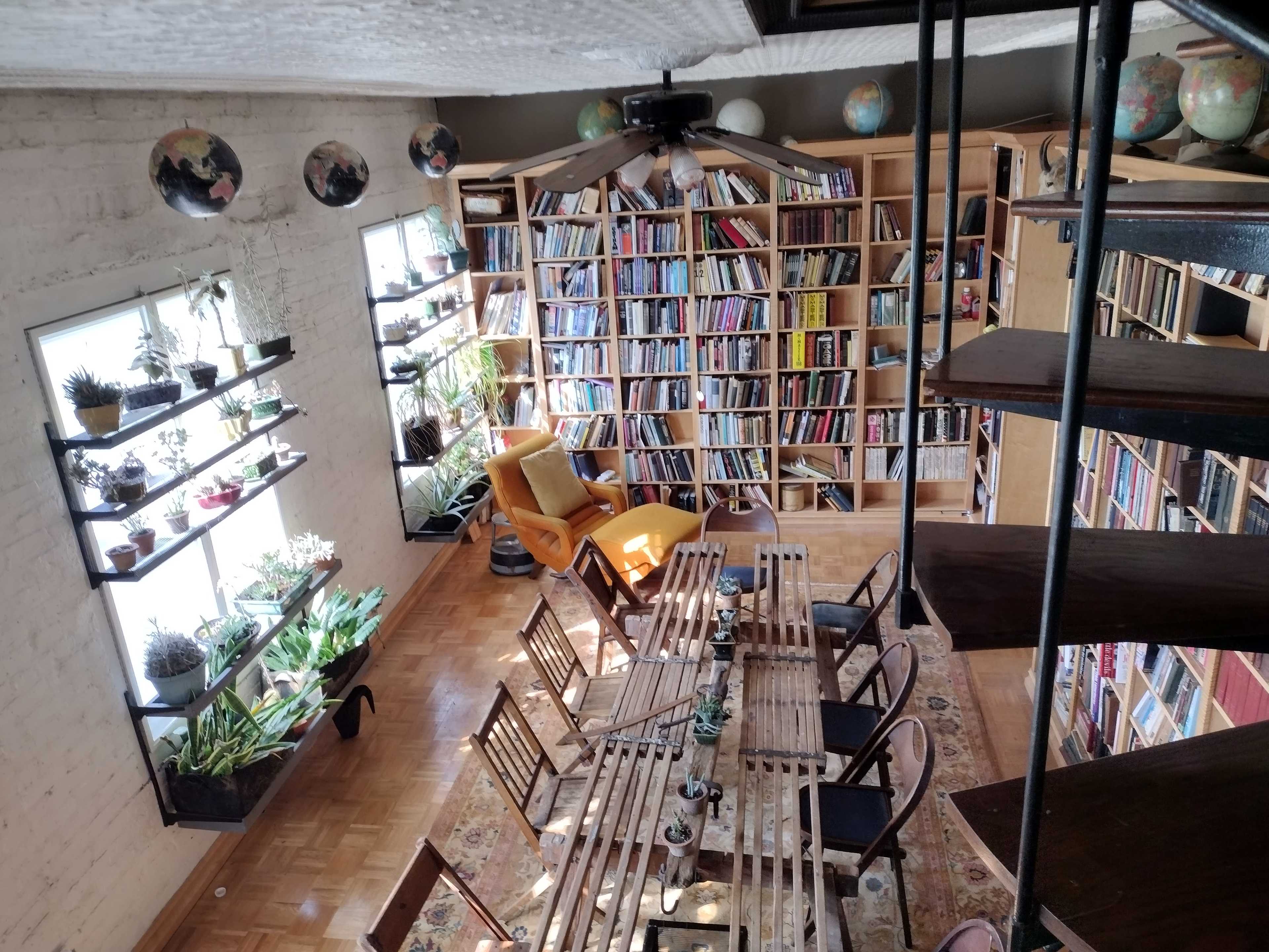 A cozy library with wooden bookshelves filled with books, a dining table surrounded by chairs, and potted plants along the windows and shelves.
