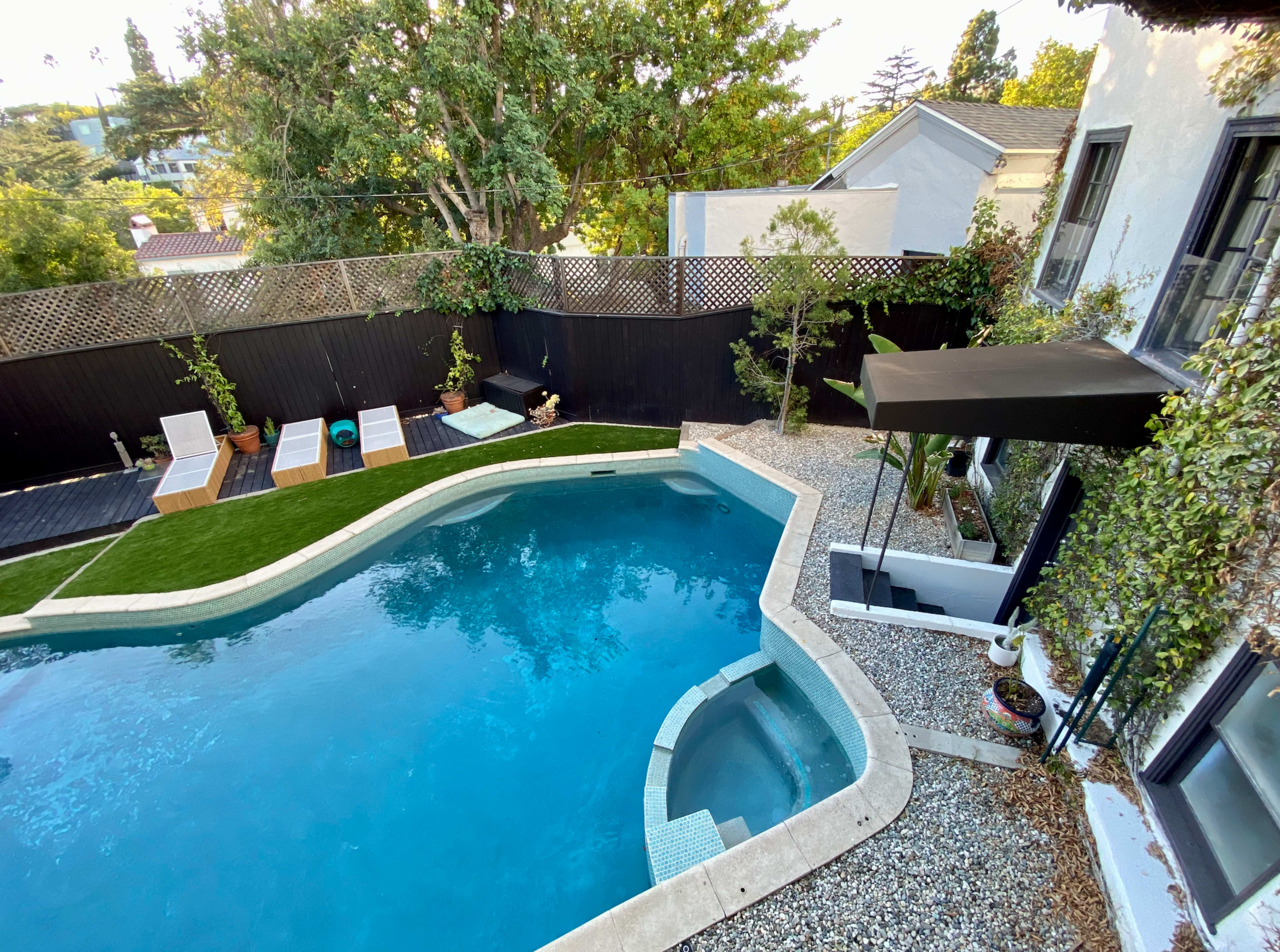 A backyard with a curved swimming pool surrounded by lounge chairs, a small garden, and a black fence.