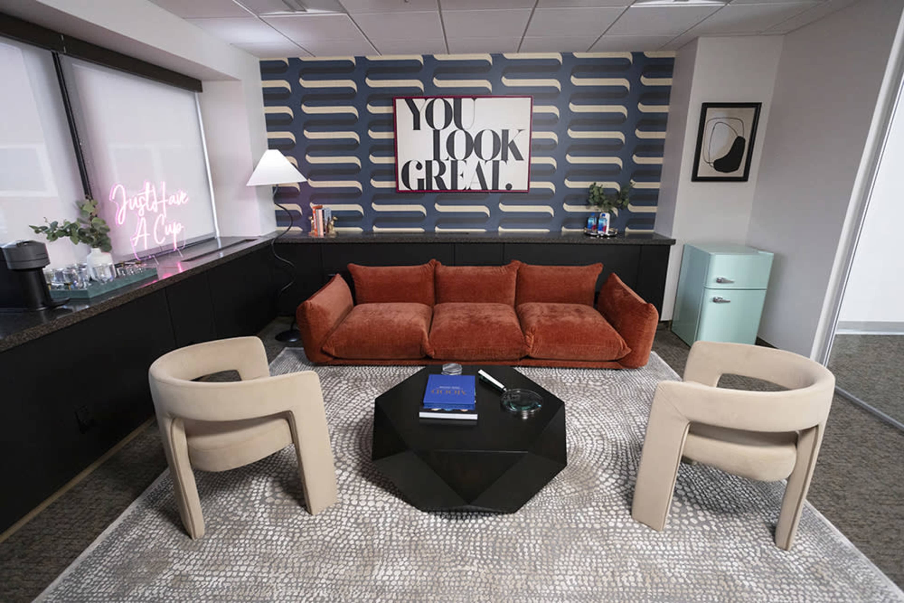 The image shows a modern office lounge area with an orange couch, two beige chairs, a black coffee table, and a patterned wall behind the seating arrangement.