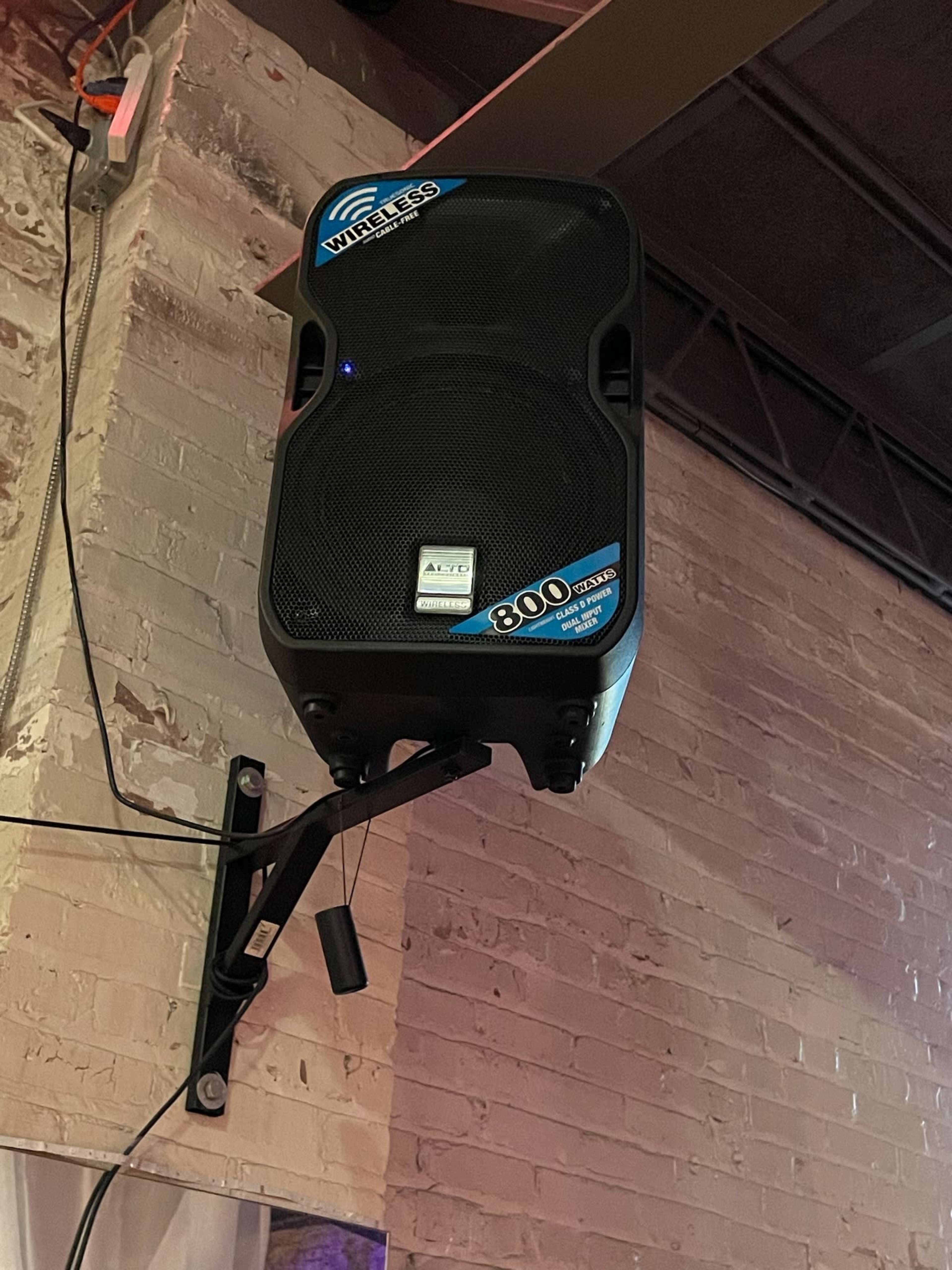 A wireless speaker is mounted on a wall with visible cables and a textured brick background.
