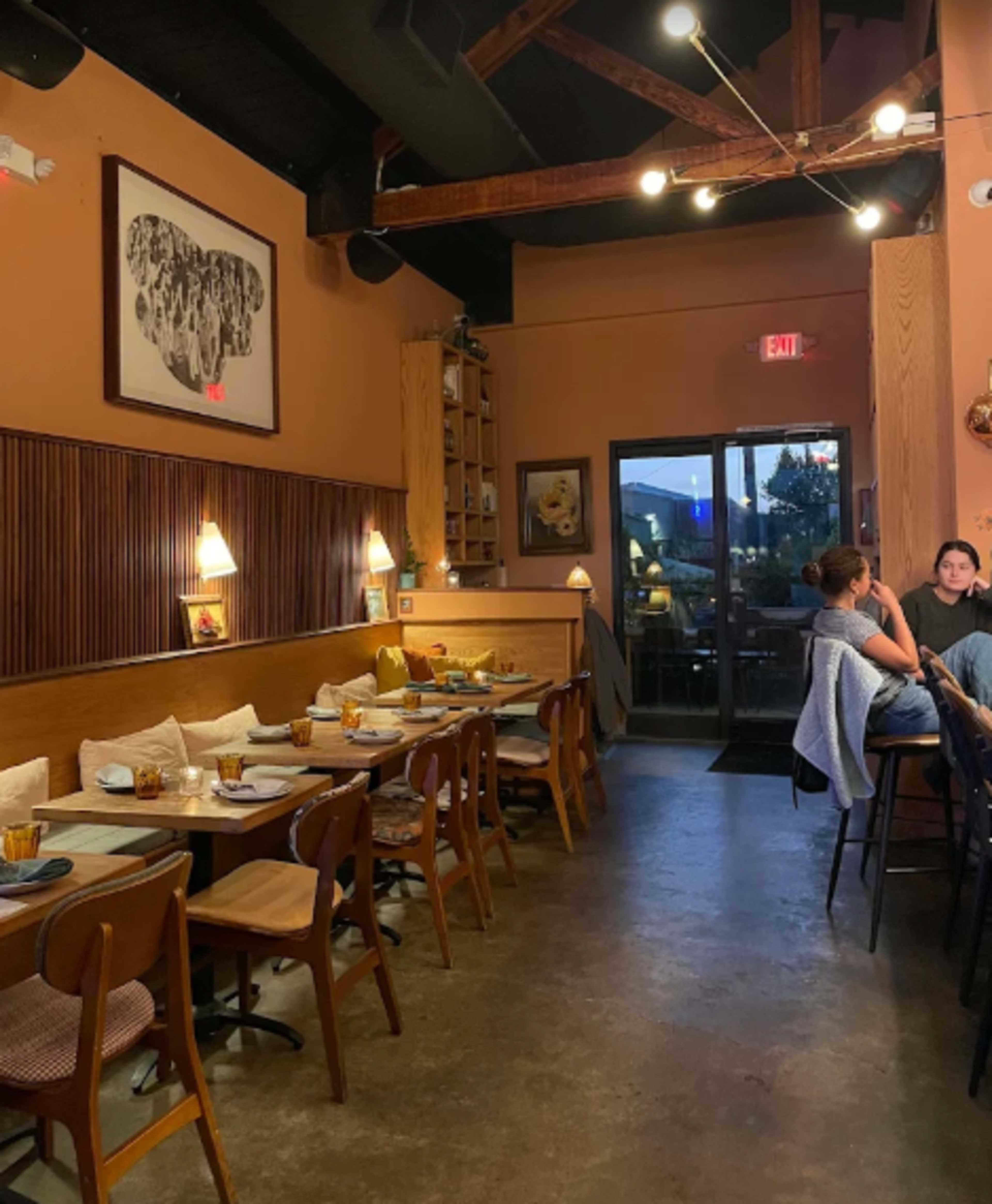 The image shows the interior of a restaurant featuring wooden accents, tables set for dining, and a large artwork on the wall.