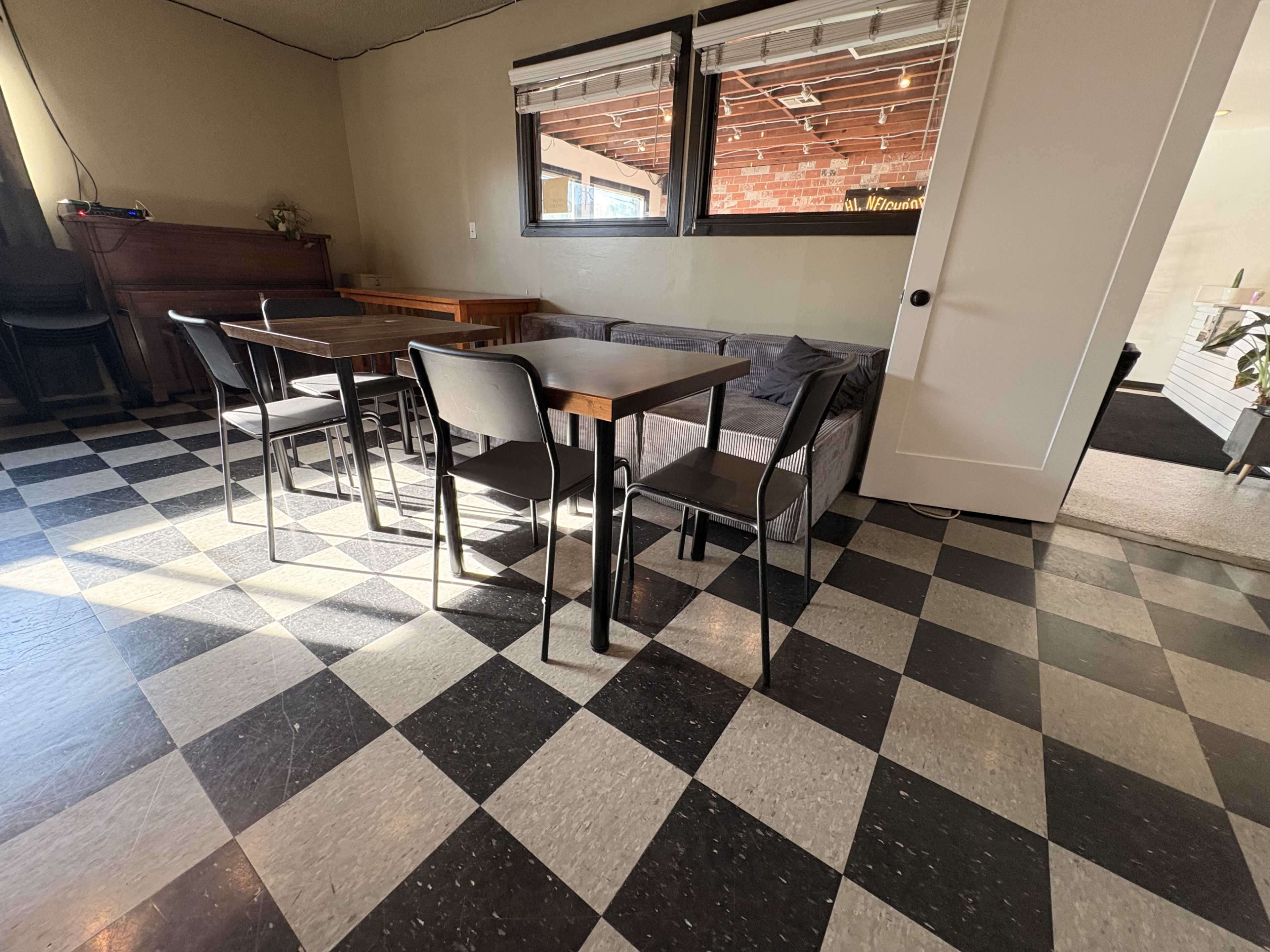 The image shows a room with checkered black and white flooring, featuring several tables and chairs, along with a sofa and large windows.