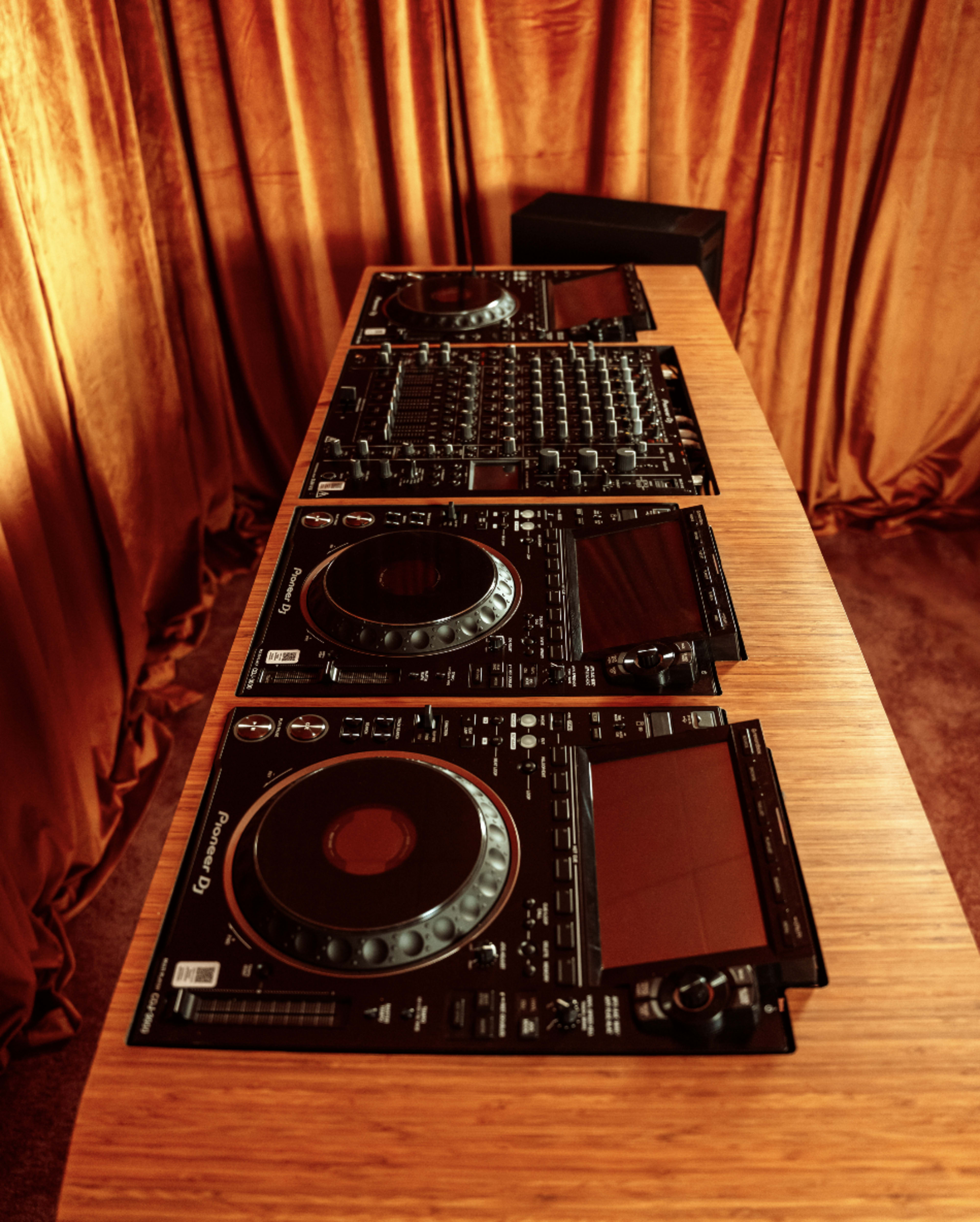 A row of professional DJ equipment is arranged neatly on a wooden table, set against a backdrop of orange curtains.