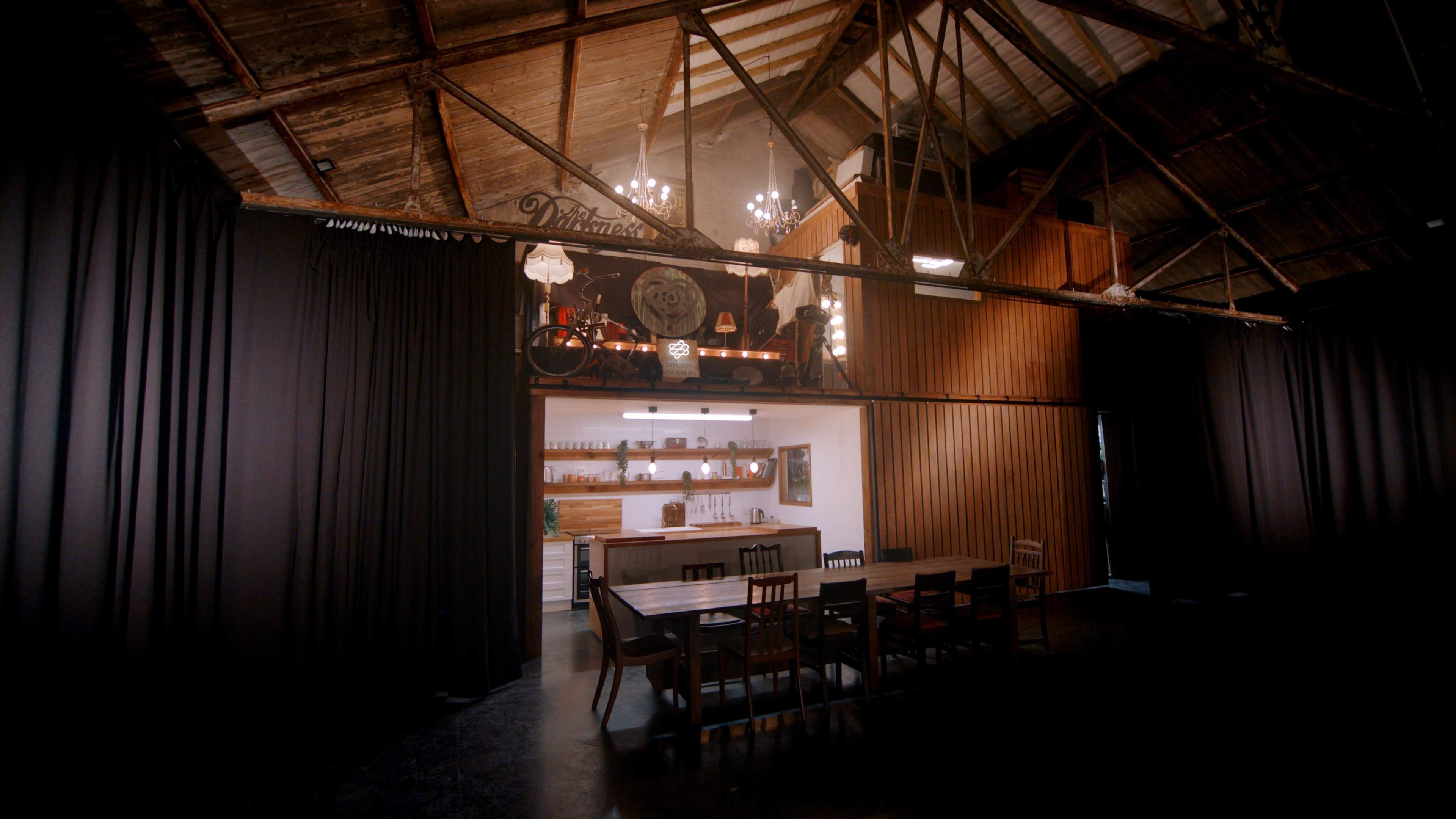 The image depicts a spacious, industrial-style dining area with a long table and wooden accents, illuminated by chandeliers, leading to an open kitchen in the background.