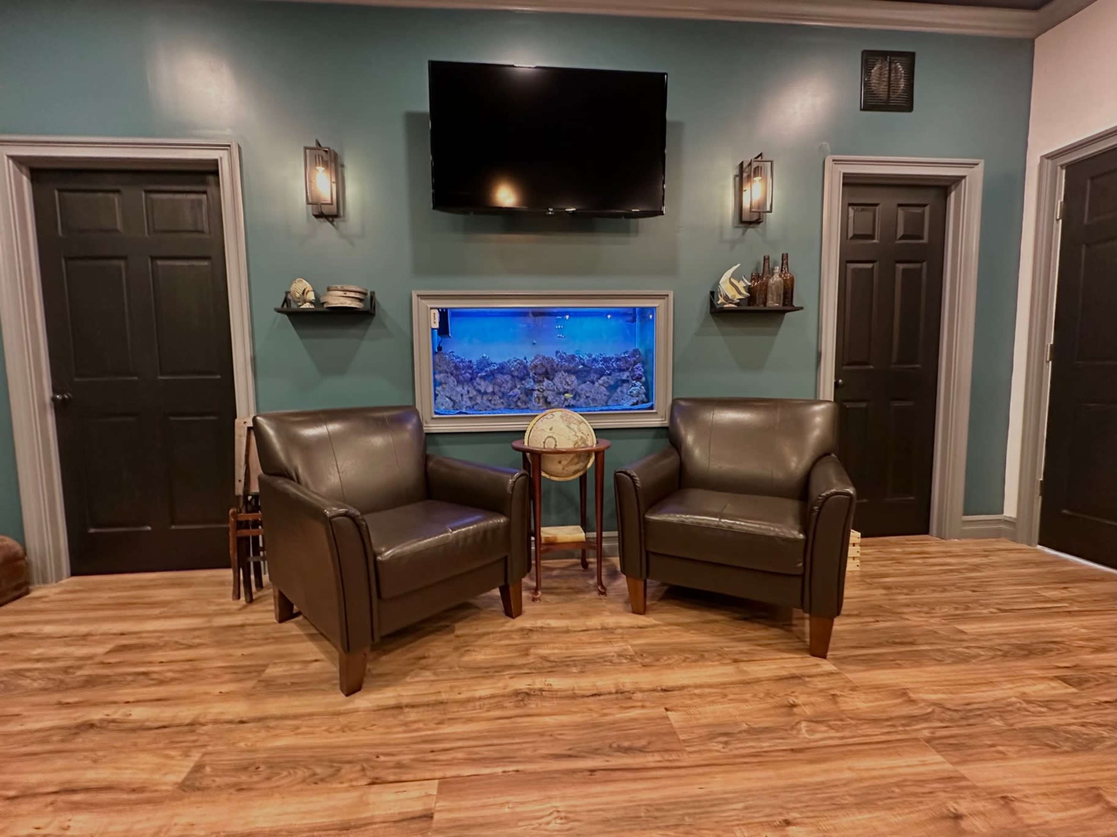 The image shows a sitting area with two leather chairs facing each other, a wooden table between them, and an aquarium with blue lighting on the wall.
