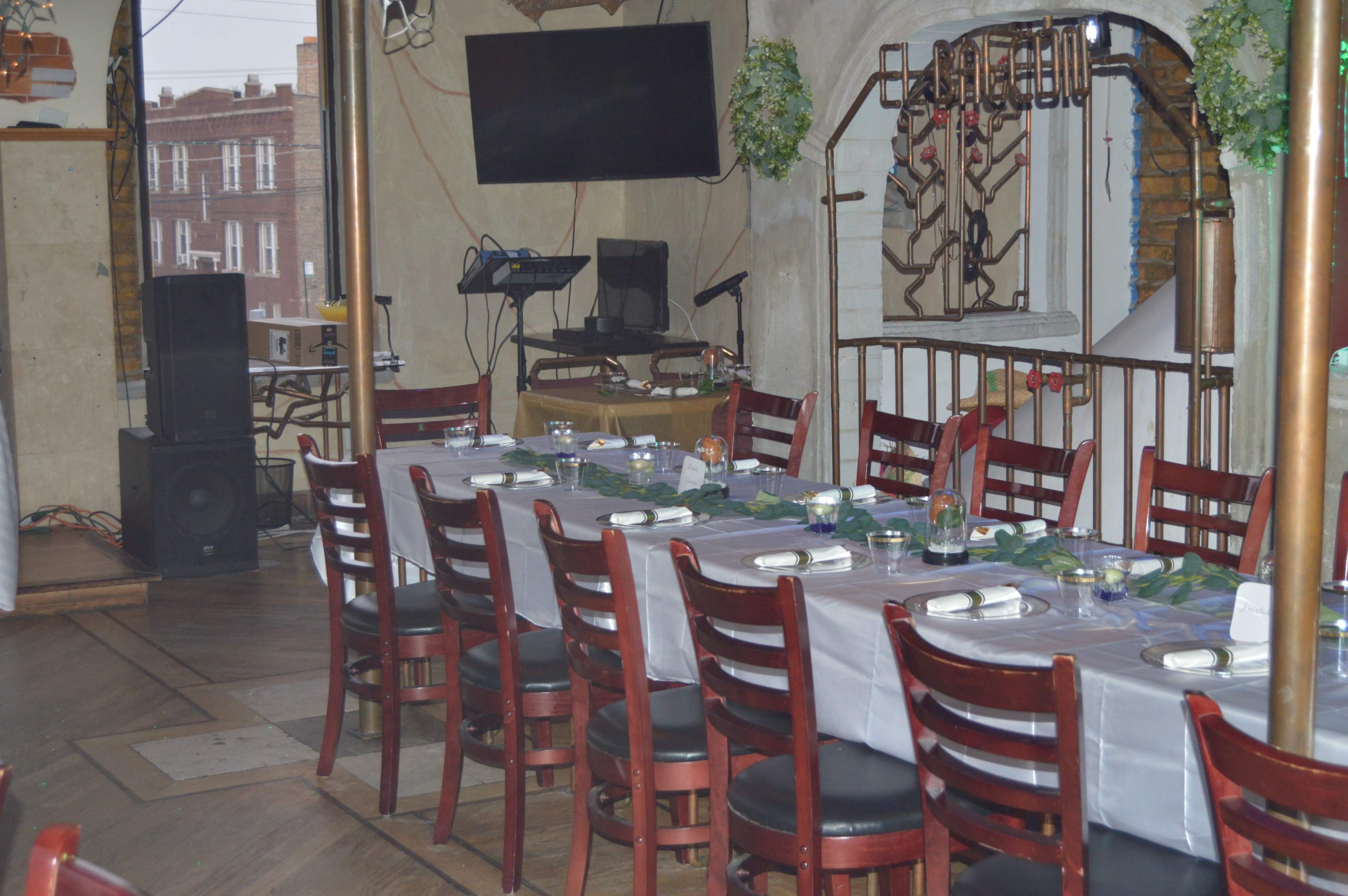A long table set with place settings and floral arrangements is positioned in a restaurant with wooden chairs and a large television on the wall.